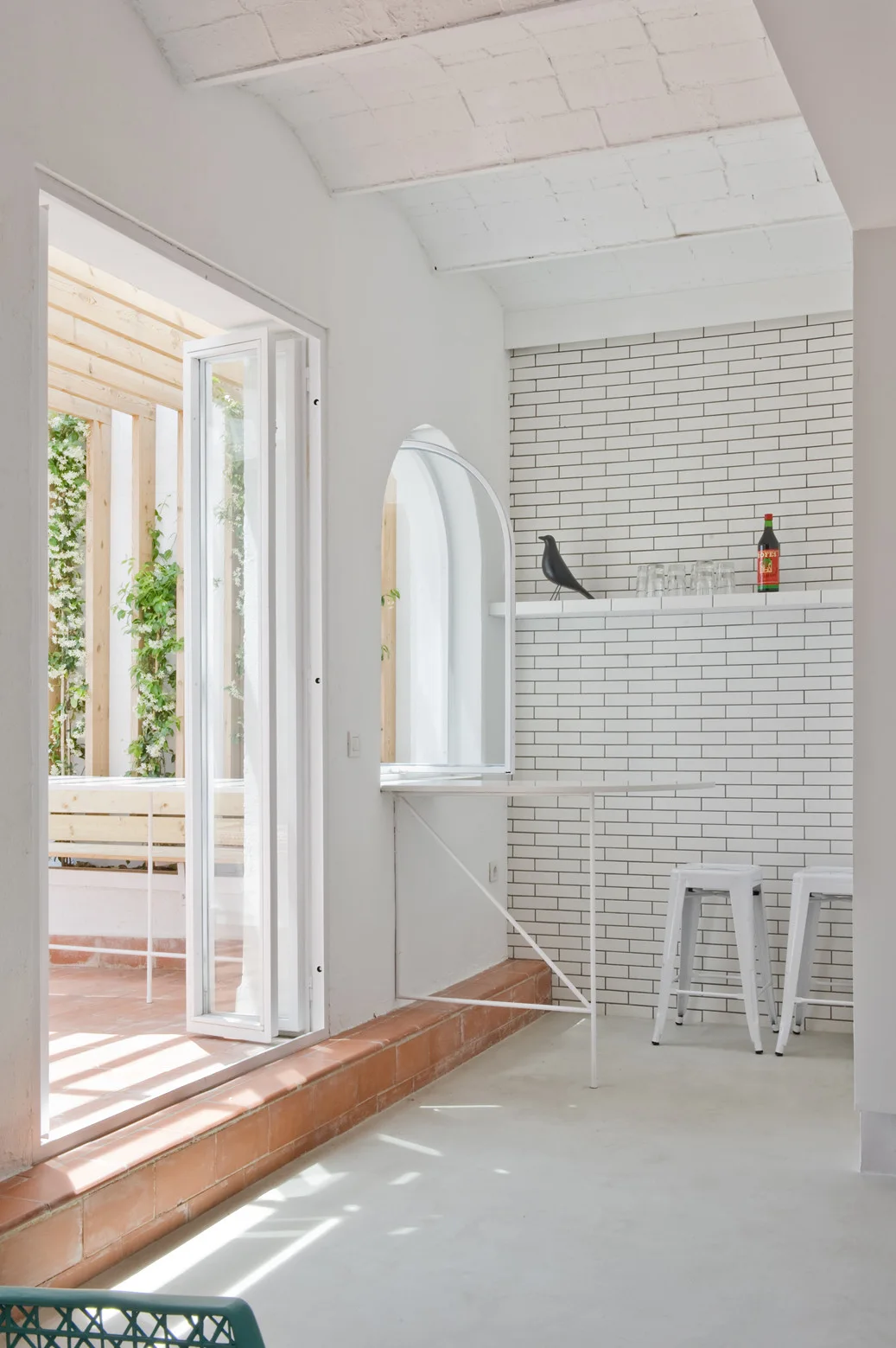 A white kitchen bar area with metal stools against a subway tile wall next to an open door leading to a terrace.