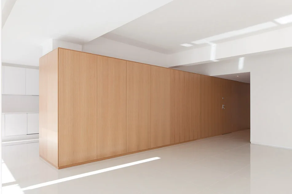 A large floor-to-ceiling light wood paneled wall partition stands in a minimalist white room with reflective tiled floors and cast shadows.