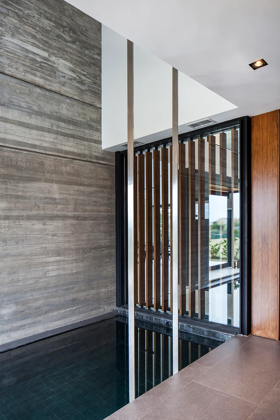An indoor water basin sits next to a textured board-formed concrete wall and a glass window featuring a vertical wooden slat screen.