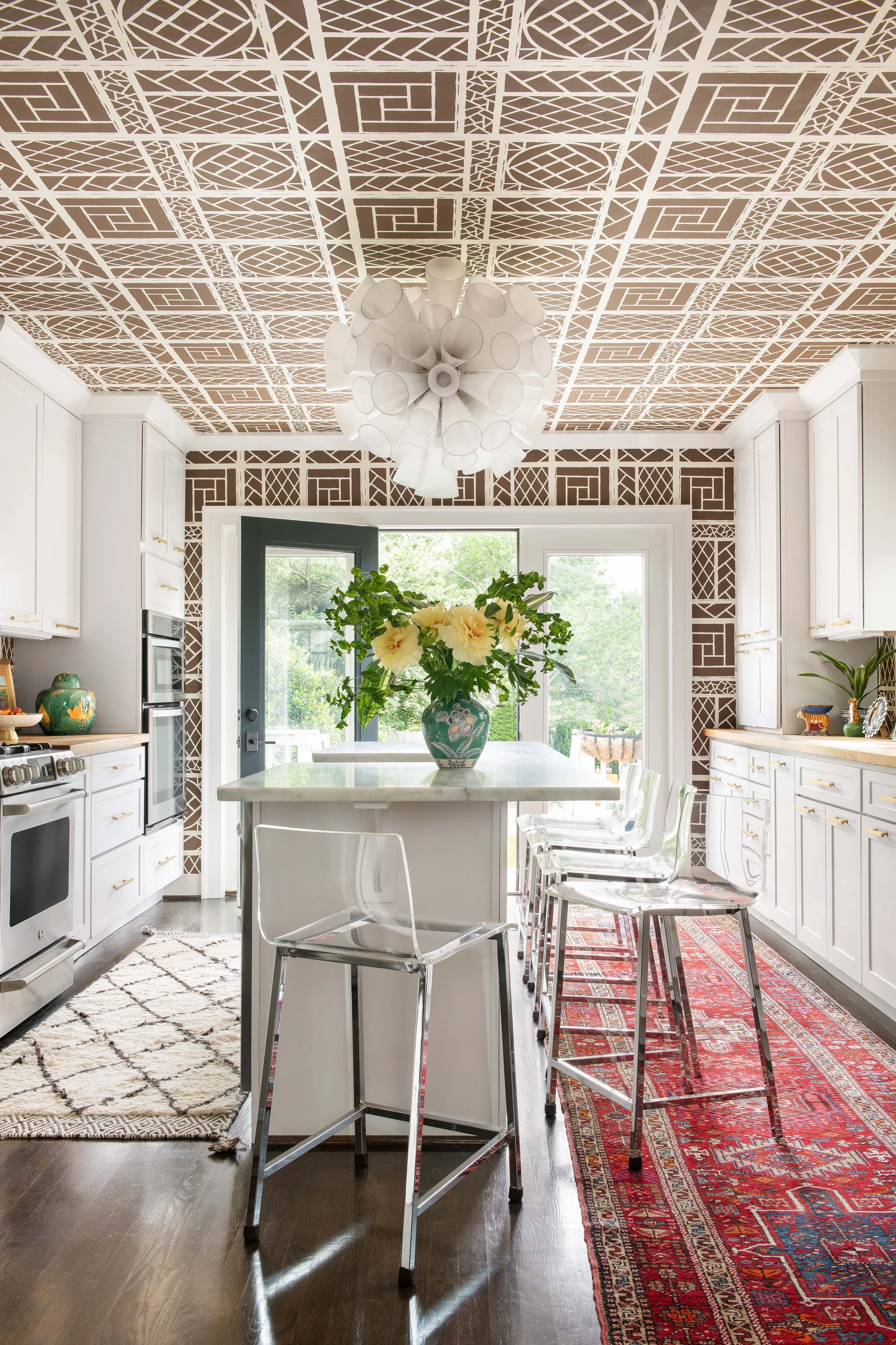 An eclectic kitchen features white shaker cabinets, a brown geometric patterned ceiling, a central marble island, and clear acrylic bar stools.