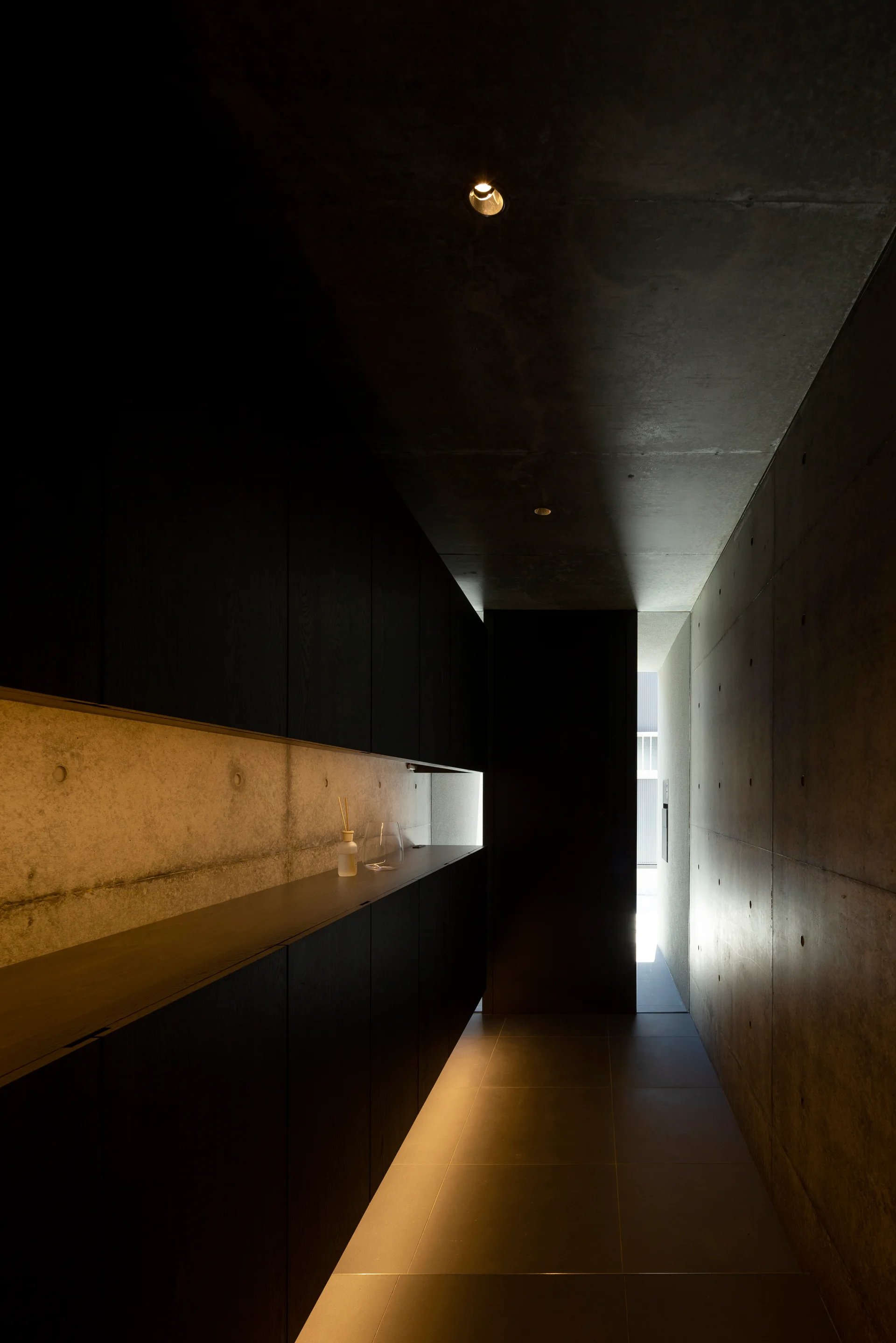 A narrow corridor with raw concrete walls, matte dark-stained built-in cabinets, and warm recessed horizontal accent lighting.