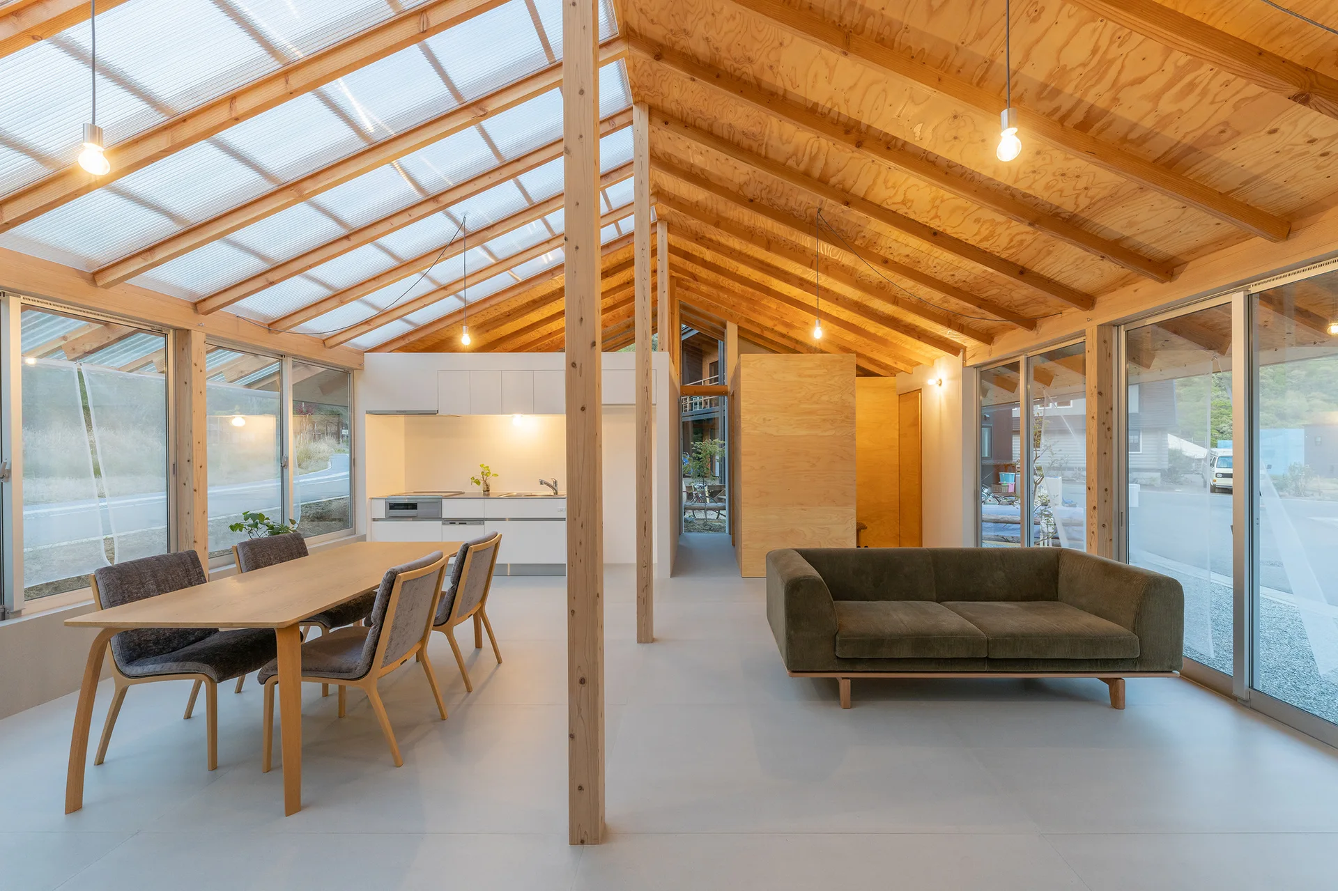 An open-plan living and dining area featuring a vaulted plywood ceiling, translucent skylight panels, a green sofa, and a wooden dining table.