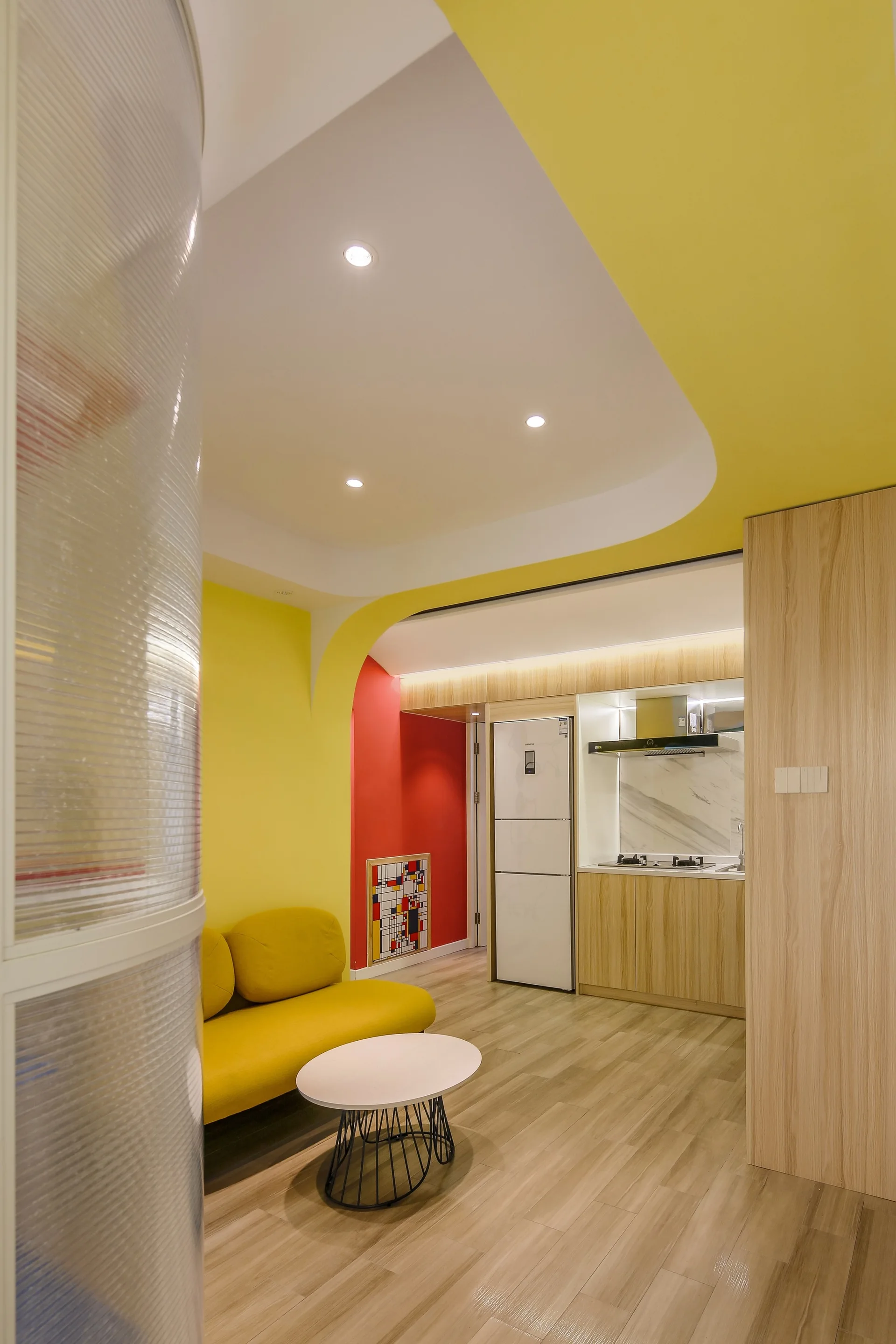 A modern open interior featuring a yellow sofa, a white round coffee table, a curved fluted glass partition, and a wood-toned kitchenette.
