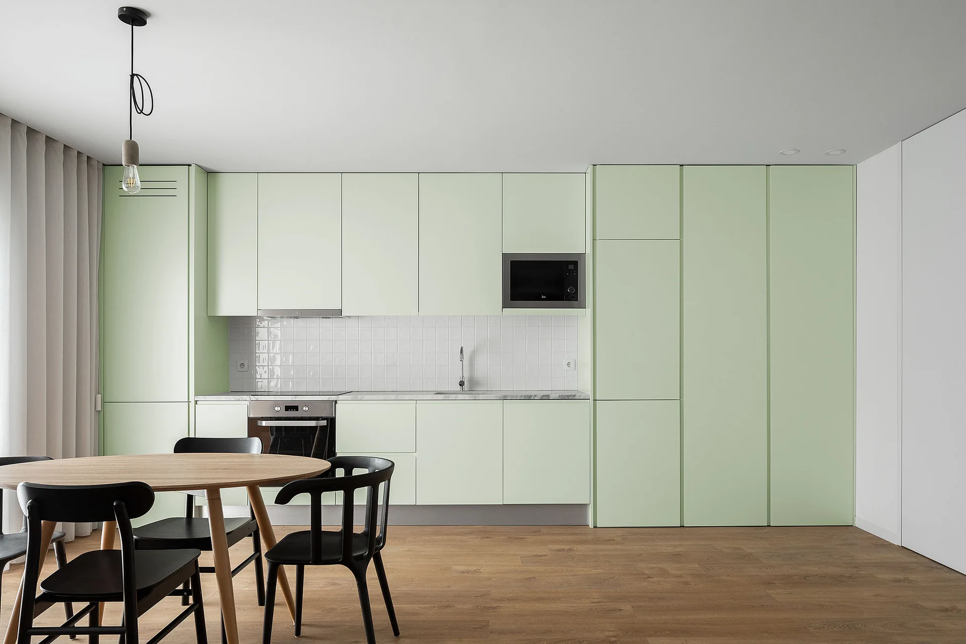 A modern minimalist kitchen features pale green flat-panel cabinets, a white tiled backsplash, wood flooring, and a round dining table with black chairs.