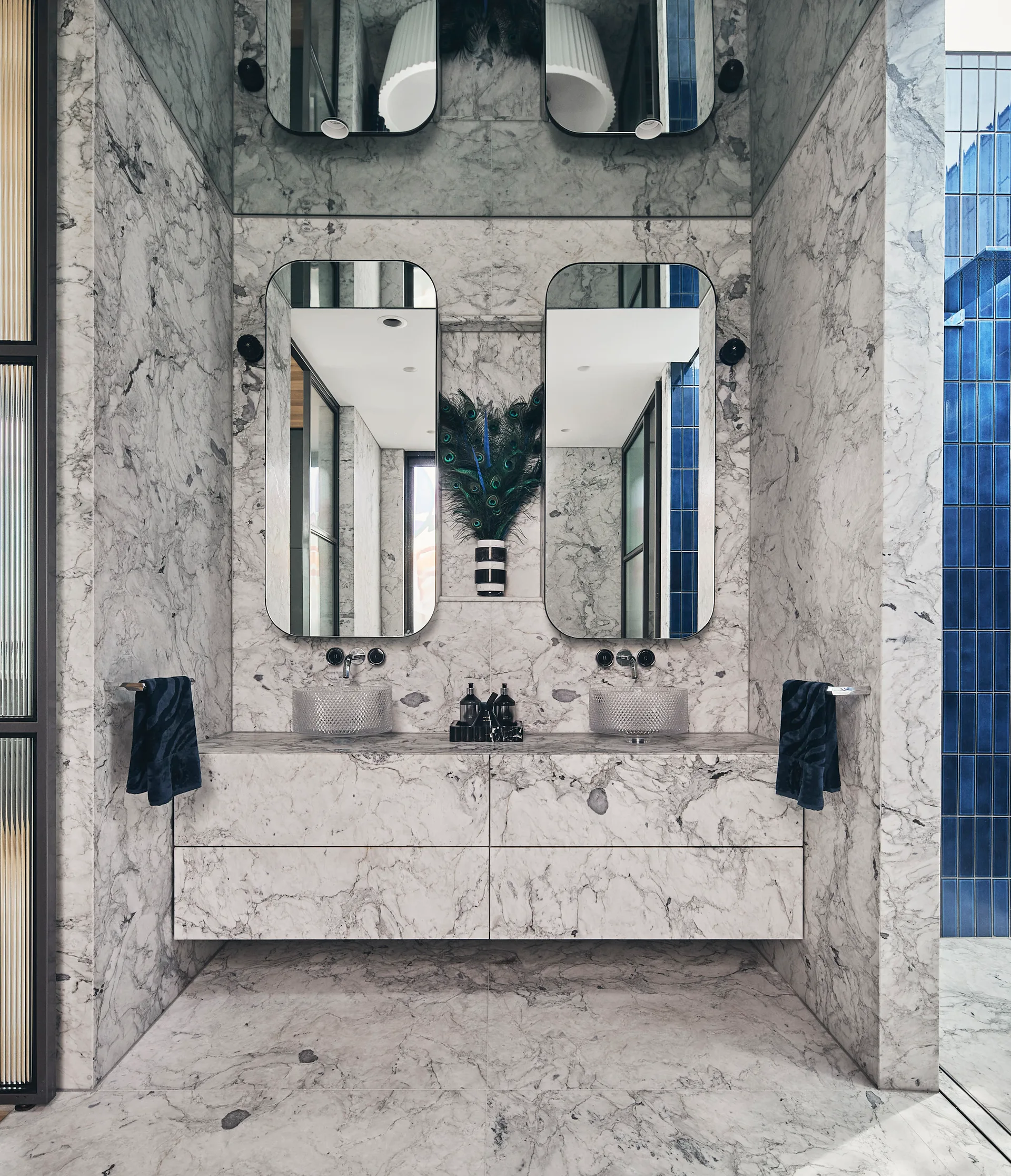 A marble-clad bathroom featuring a floating stone vanity with textured glass vessel sinks, two arched mirrors, and a striped vase.
