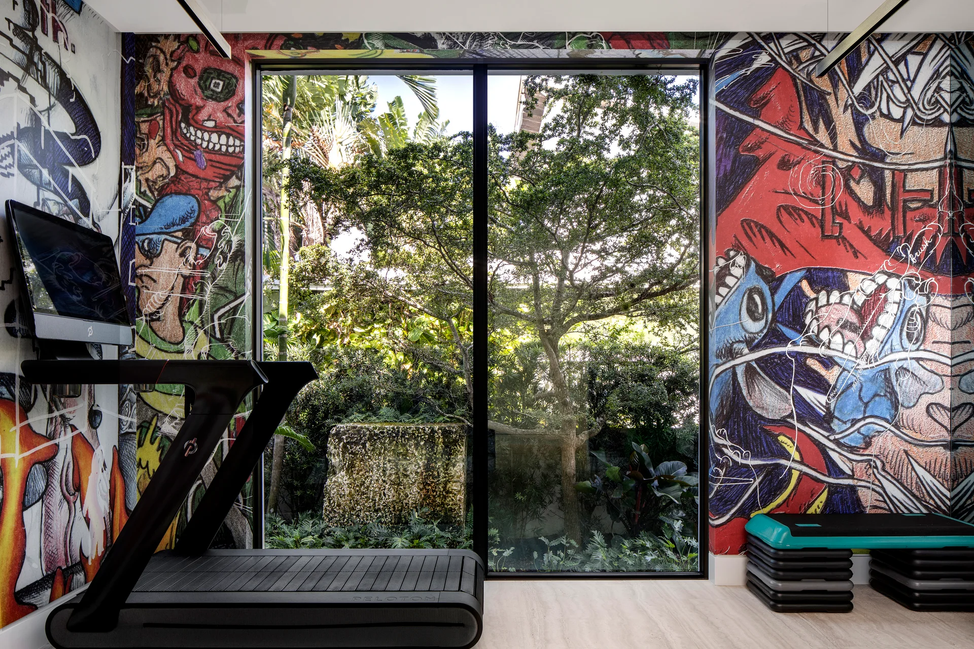 A black treadmill stands in a home gym next to stacked fitness steps, framed by colorful graffiti wallpaper and a large window looking out at a garden.