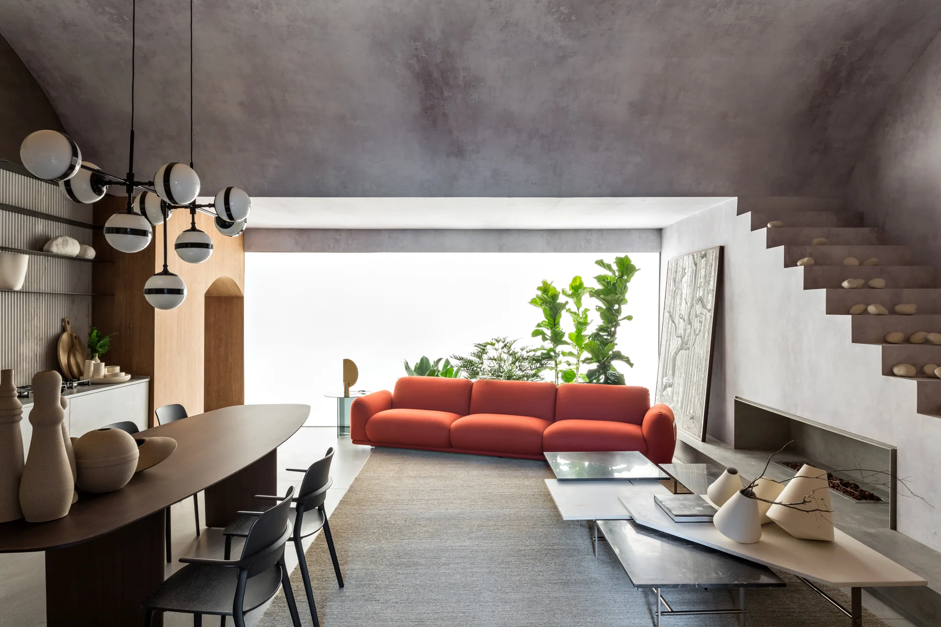 A modern open-plan living room features a bright red sofa, an elliptical dark wood dining table, an orb chandelier, and an integrated floating staircase.