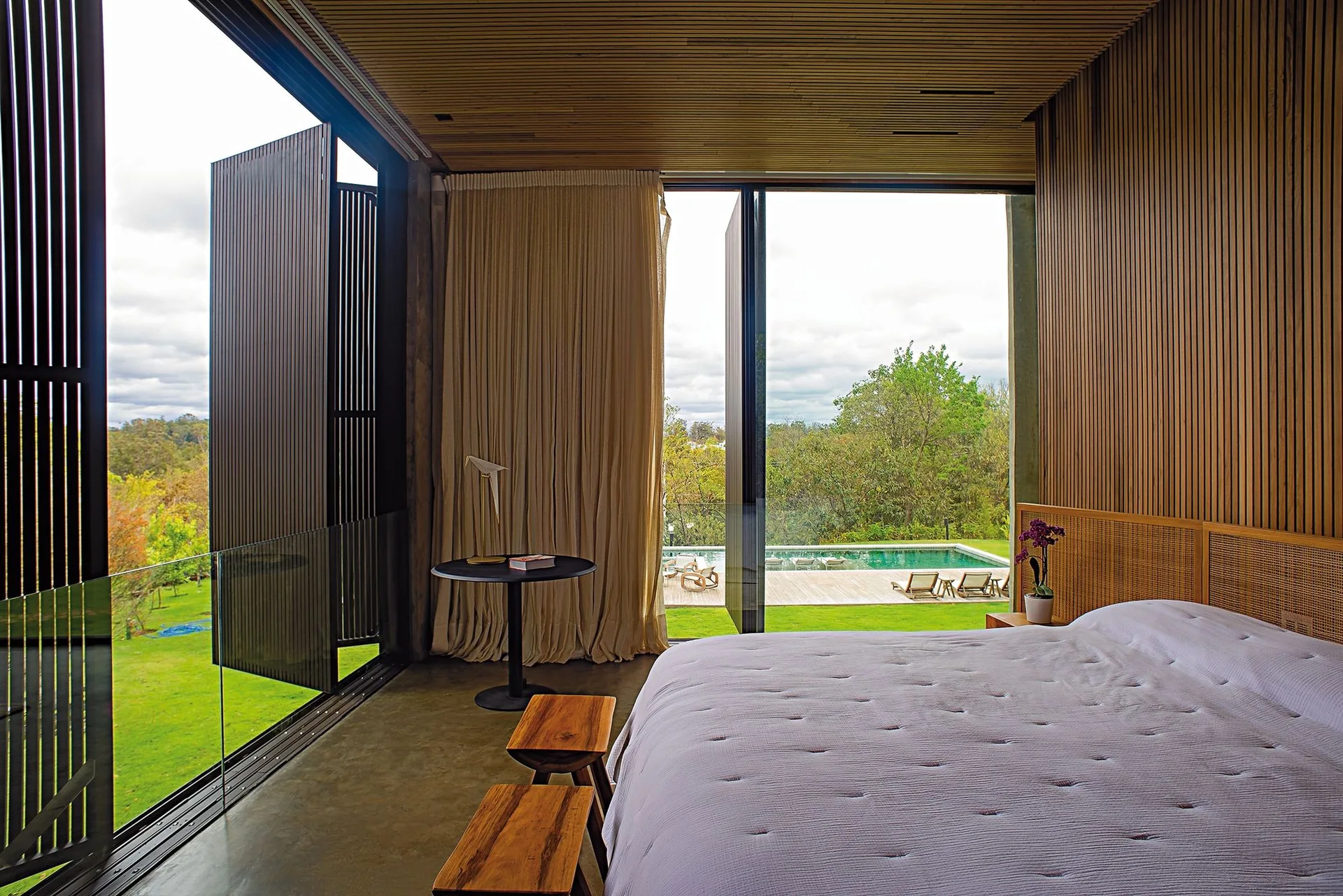 A modern bedroom features wood-slat walls and ceiling, a bed with a woven headboard, and large glass doors opening to an outdoor pool.