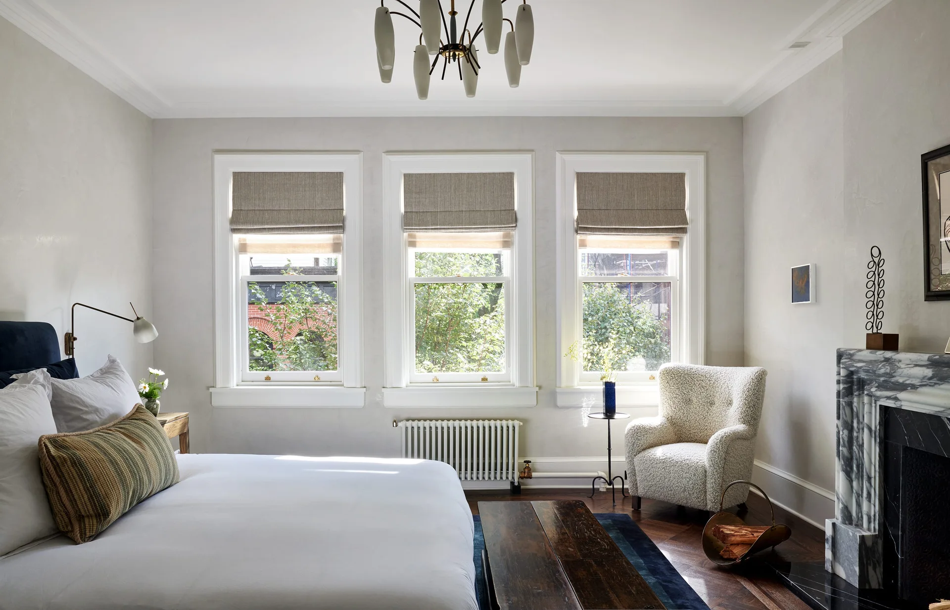 A light gray bedroom features three windows with roman shades, a marble fireplace, a textured armchair, and a bed with white linens.