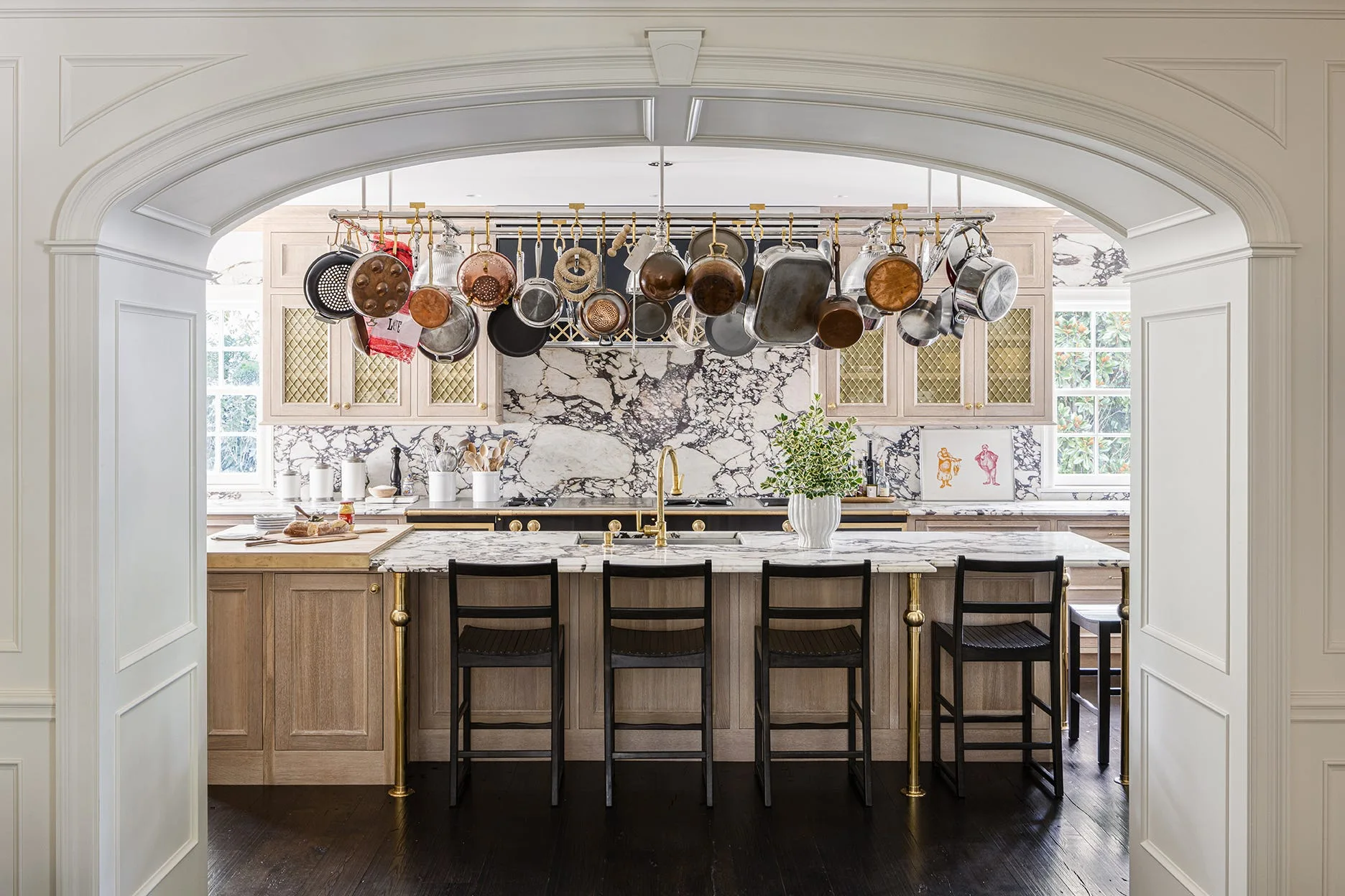 A marble kitchen island with black bar stools sits under a hanging pot rack of copper cookware, viewed through a white architectural archway.