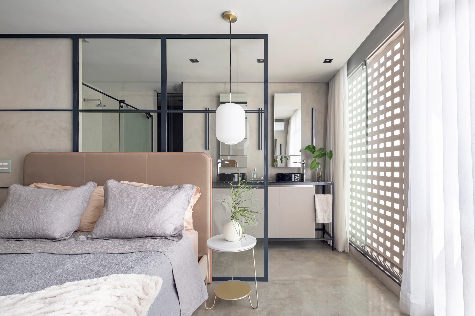 A modern bedroom with an upholstered headboard features a black-framed glass wall divider opening to an ensuite bathroom with a beige vanity.