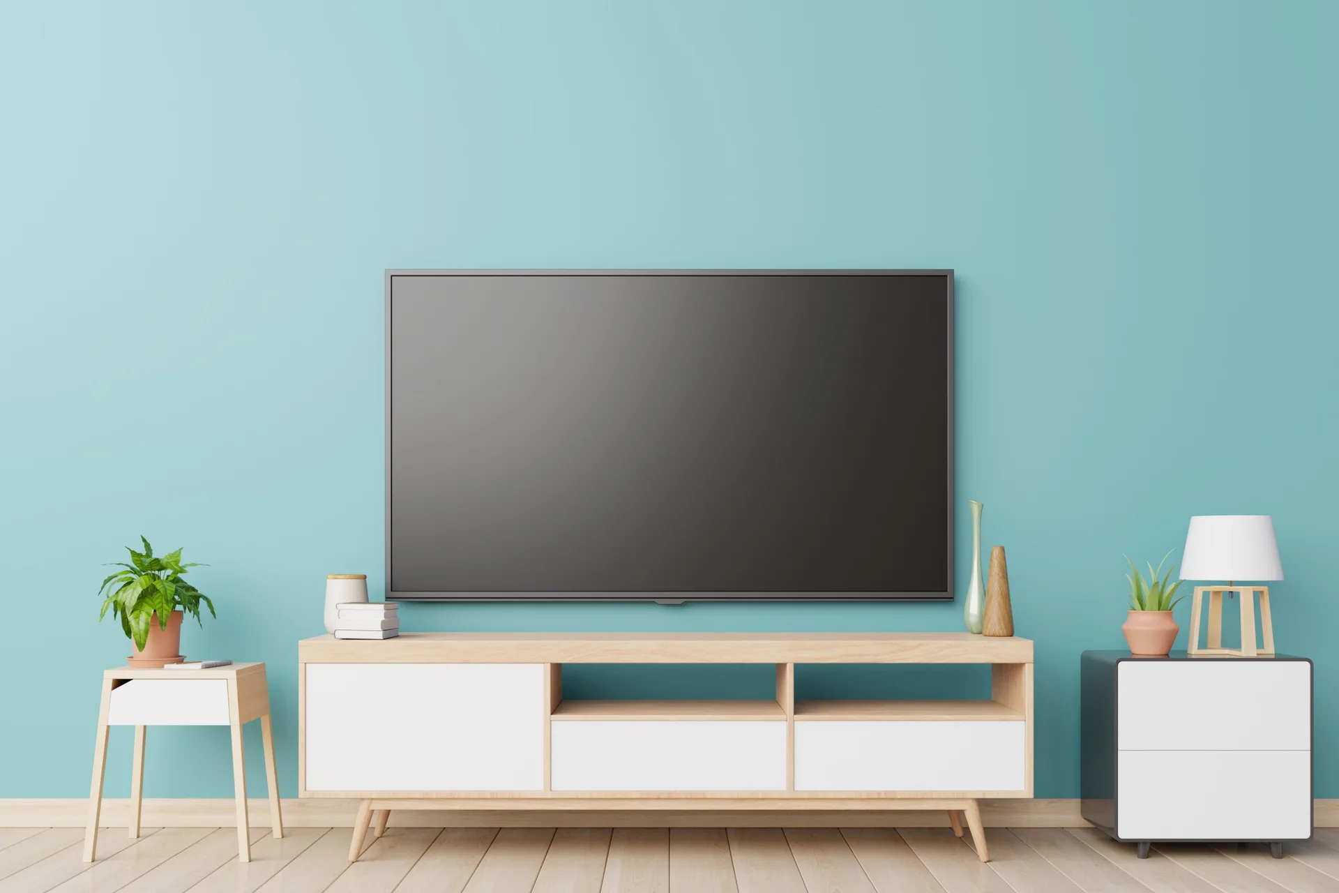 A flat-screen television is mounted on a teal wall above a light wood media console and two small accent tables.