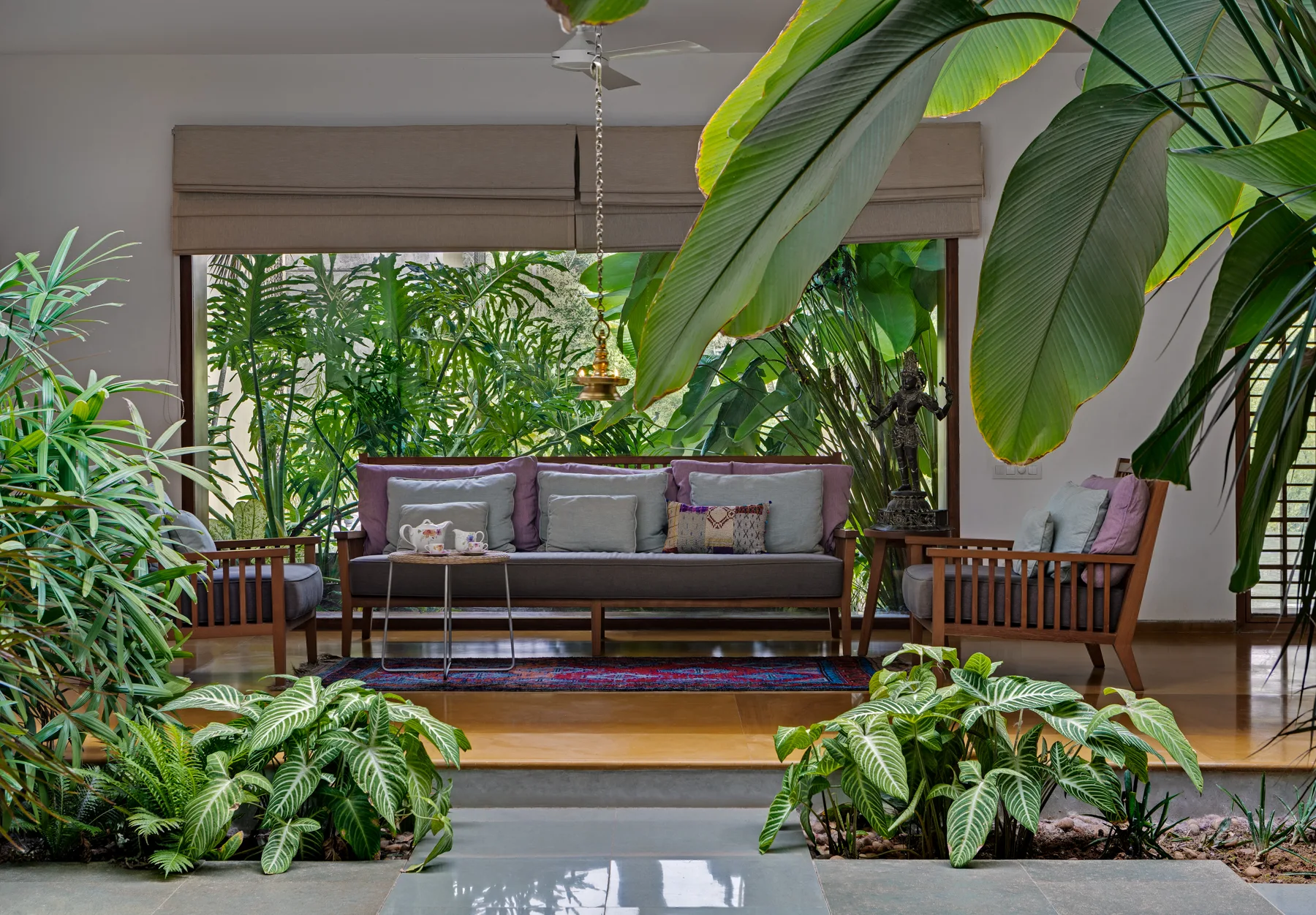 A sunken living room features a wooden sofa and armchairs surrounded by lush green plants, with a large window and roman shades in the background.