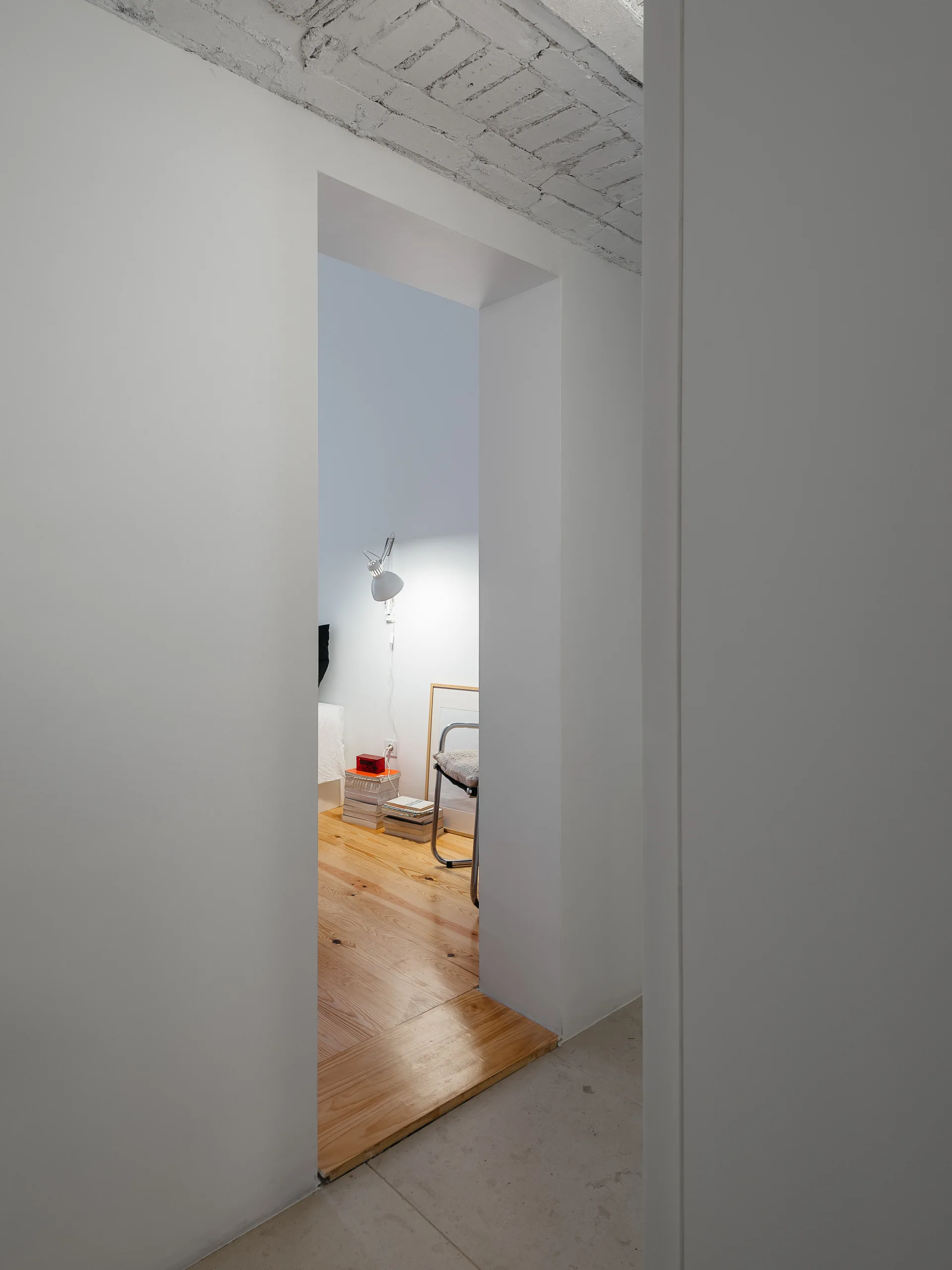 A view through a white doorway reveals a minimalist bedroom with light wood flooring, a white task light, and a painted brick ceiling.