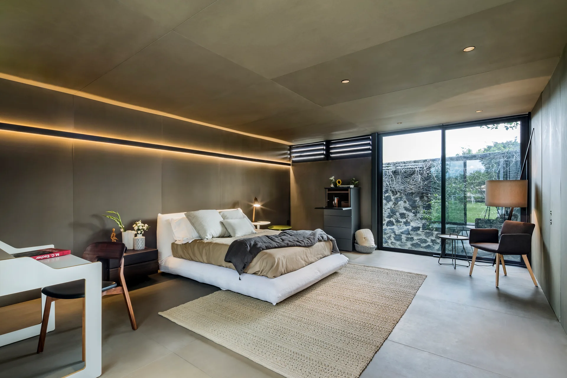 A modern minimalist bedroom featuring a low platform bed, matte dark wall paneling with a horizontal LED strip, and large glass doors opening to a garden.