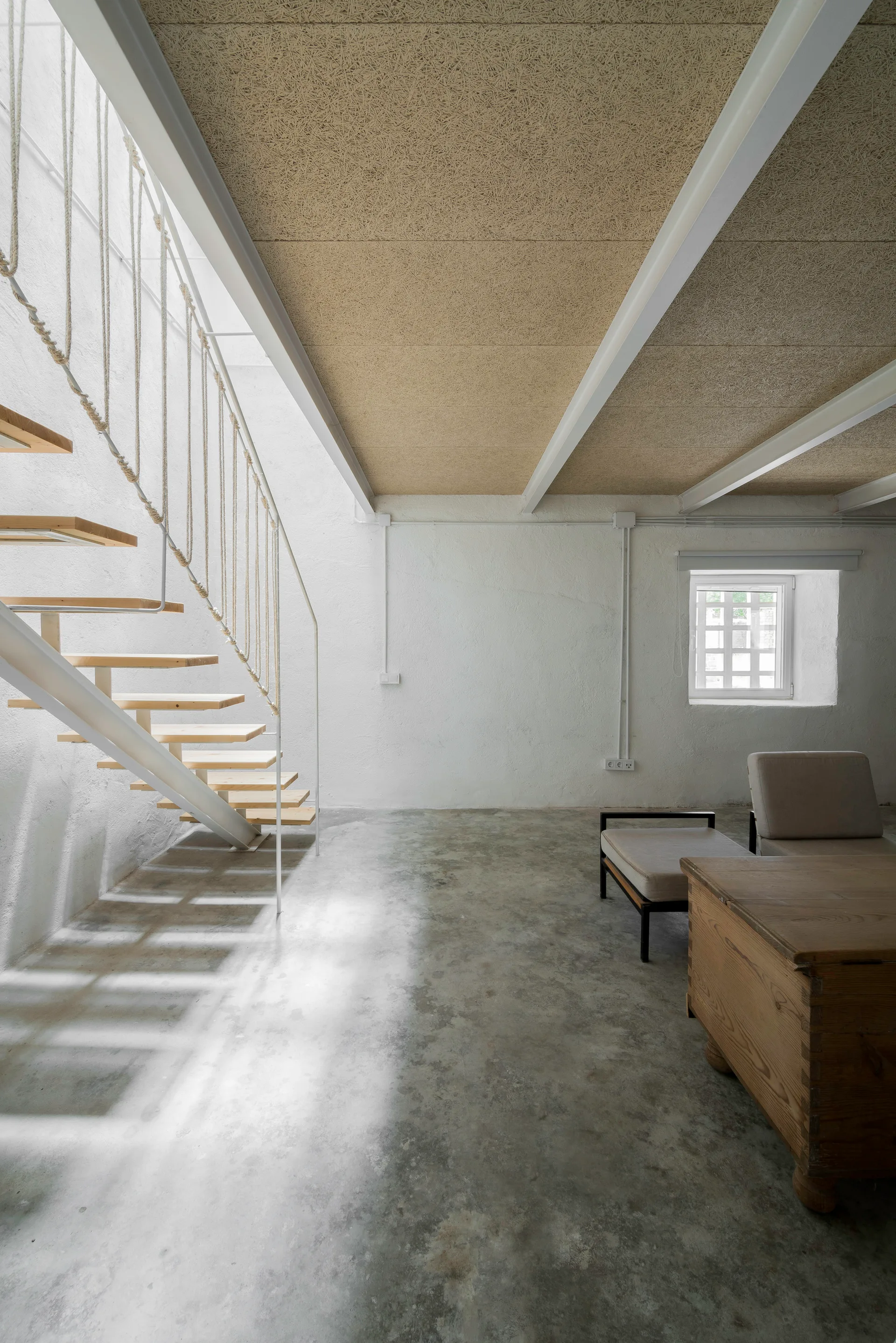 A minimalist room features an open-riser staircase with rope railings, a polished concrete floor, and textured ceiling panels.