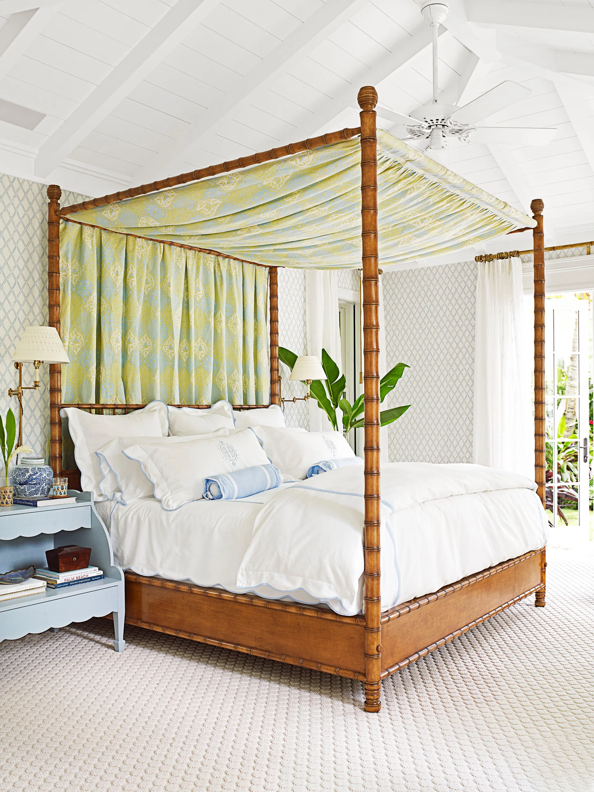 A bright bedroom features a carved wooden canopy bed draped in yellow and blue fabric, next to a blue scalloped nightstand under a white vaulted ceiling.
