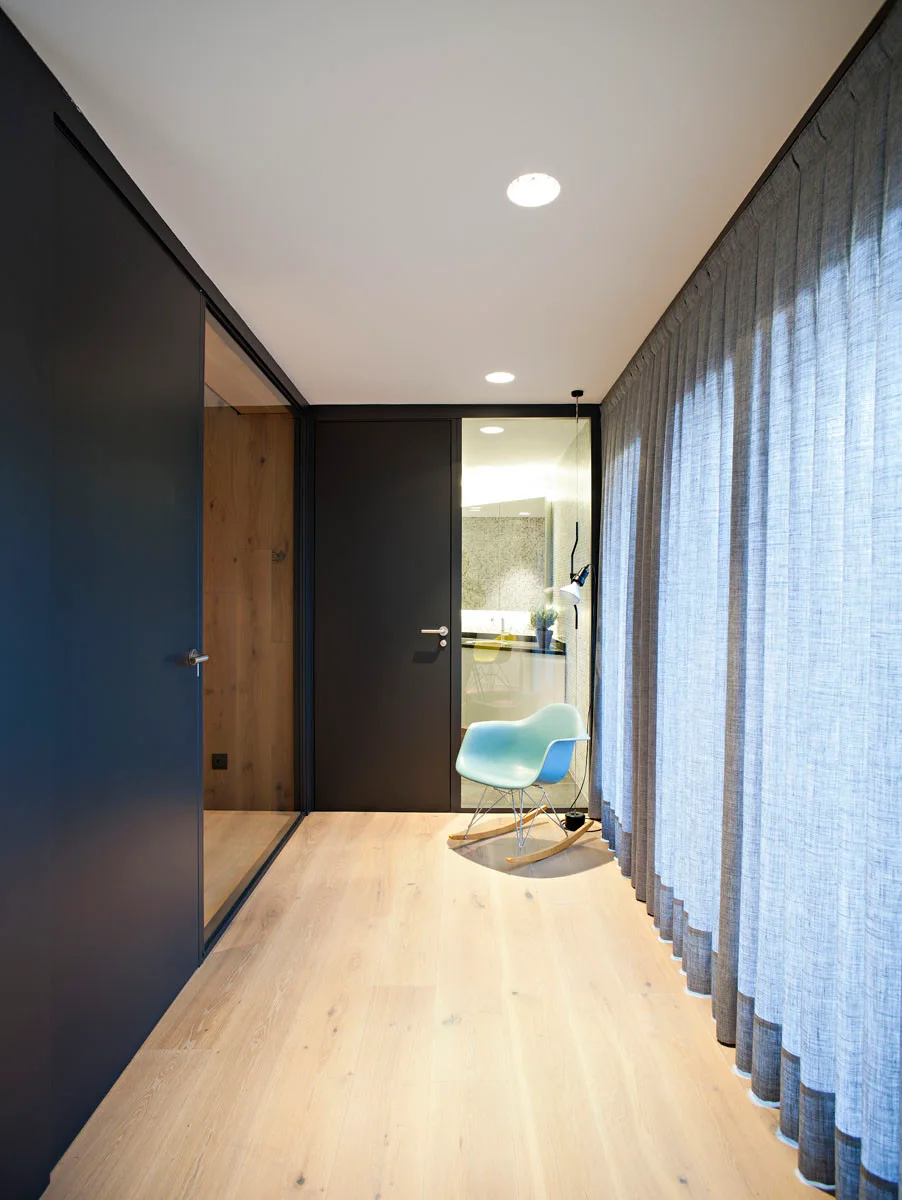 A minimalist hallway with light wood floors, dark matte doors, a full-length grey curtain, and a light blue rocking chair.