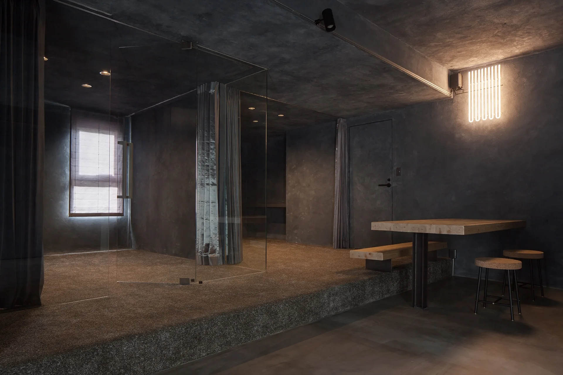 A dark gray minimalist room featuring a raised carpeted platform with a glass partition, a built-in wooden dining table, and a linear wall light.