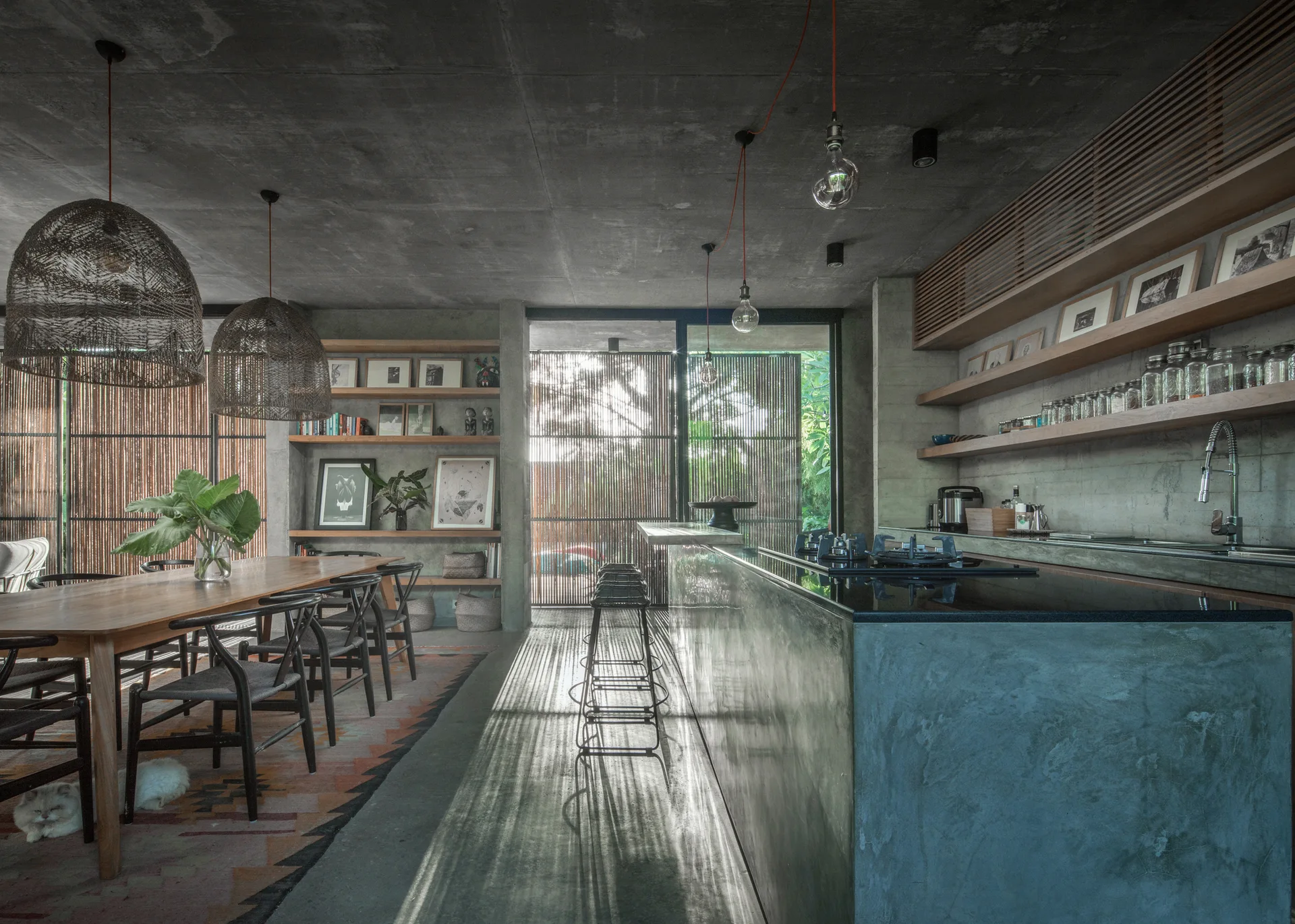 An open-concept kitchen and dining area with an exposed concrete ceiling, a concrete island, a wooden dining table, and slatted bamboo screens.
