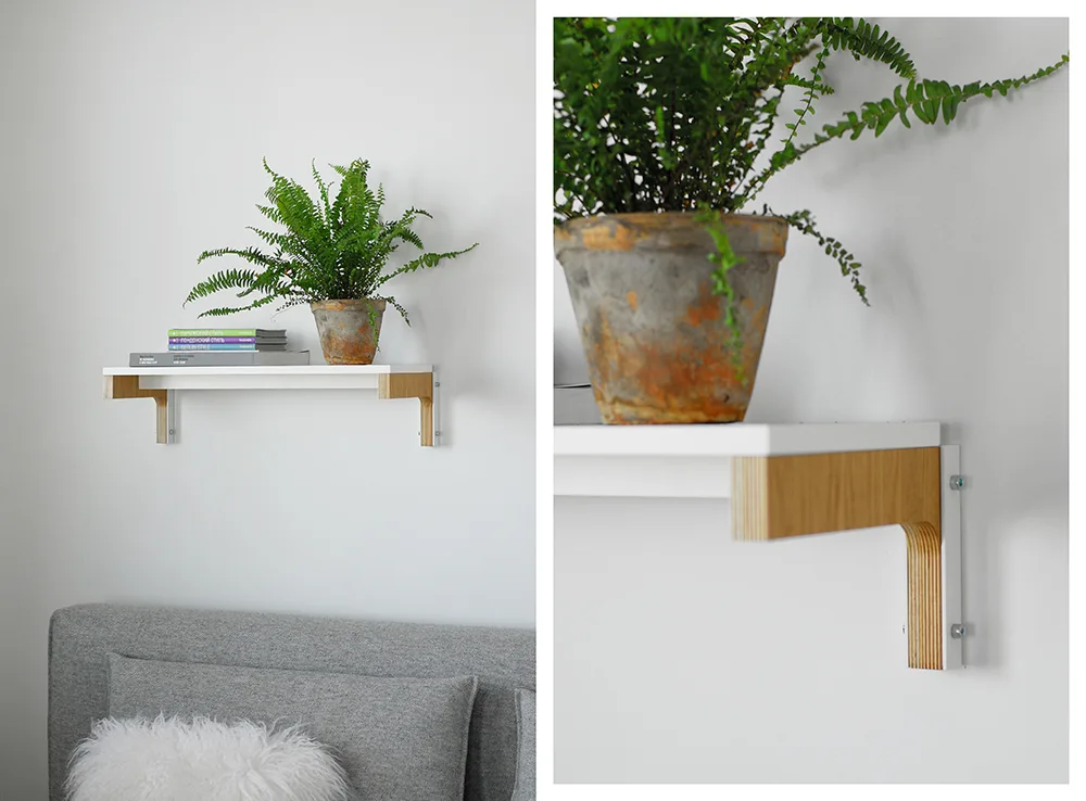 A white wall-mounted shelf with layered plywood brackets holds a potted green fern and books above a gray fabric headboard.