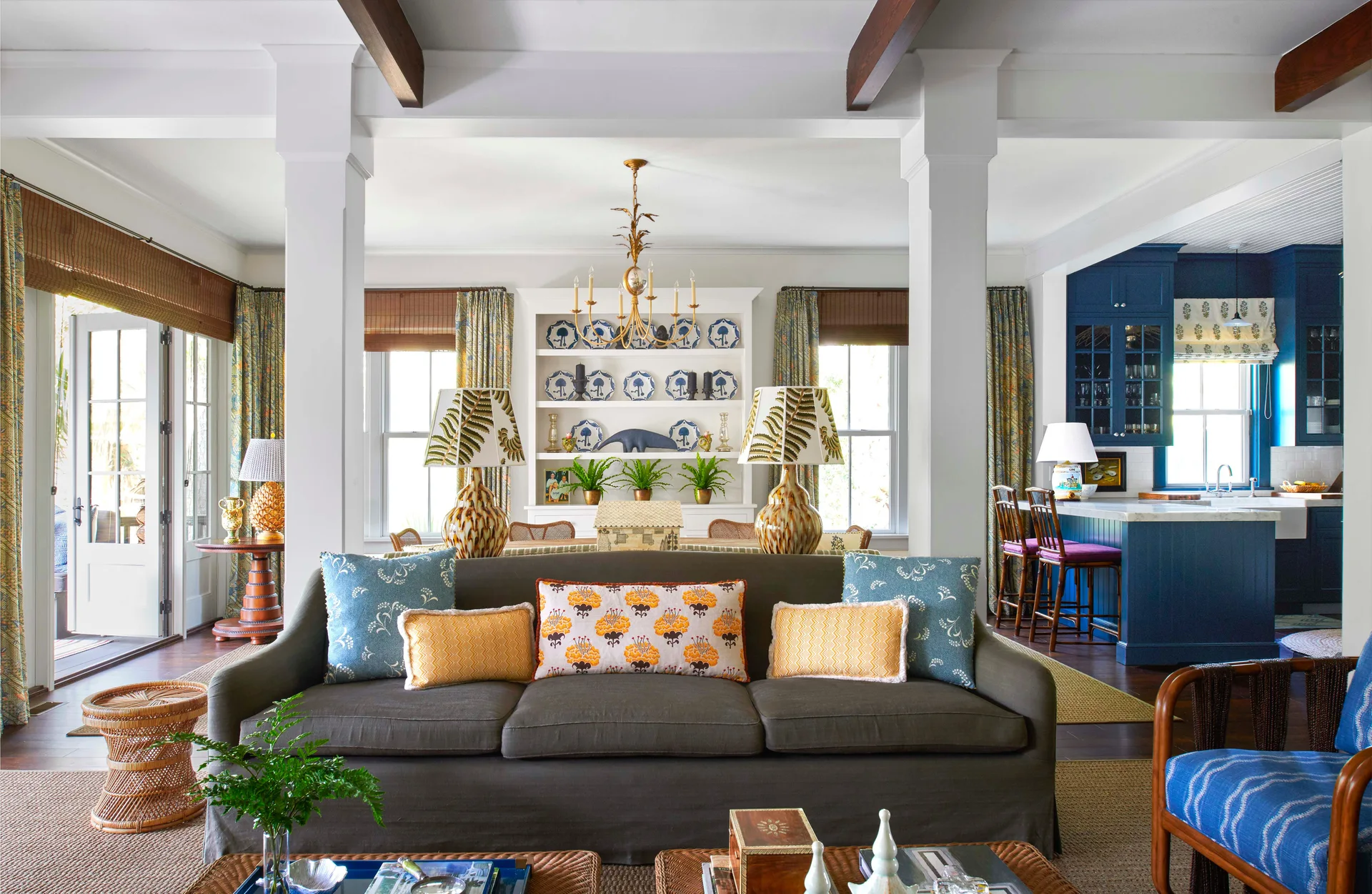 An open-concept living room featuring a grey sofa, exposed dark wood ceiling beams, white built-in shelving, and blue kitchen cabinets.
