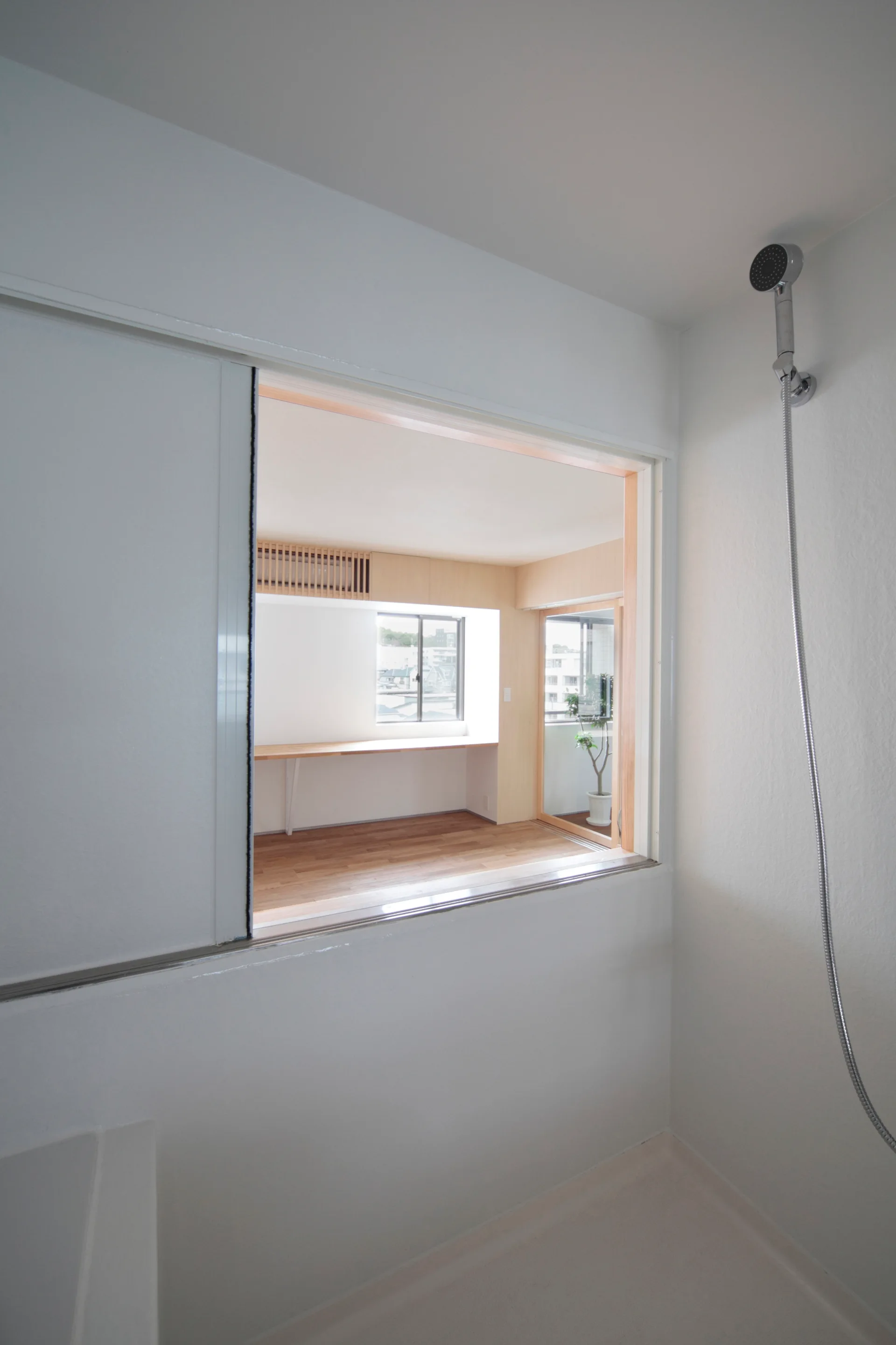 A view from a white minimalist bathroom through a large open sliding window into a sunny room with wood floors and a desk.