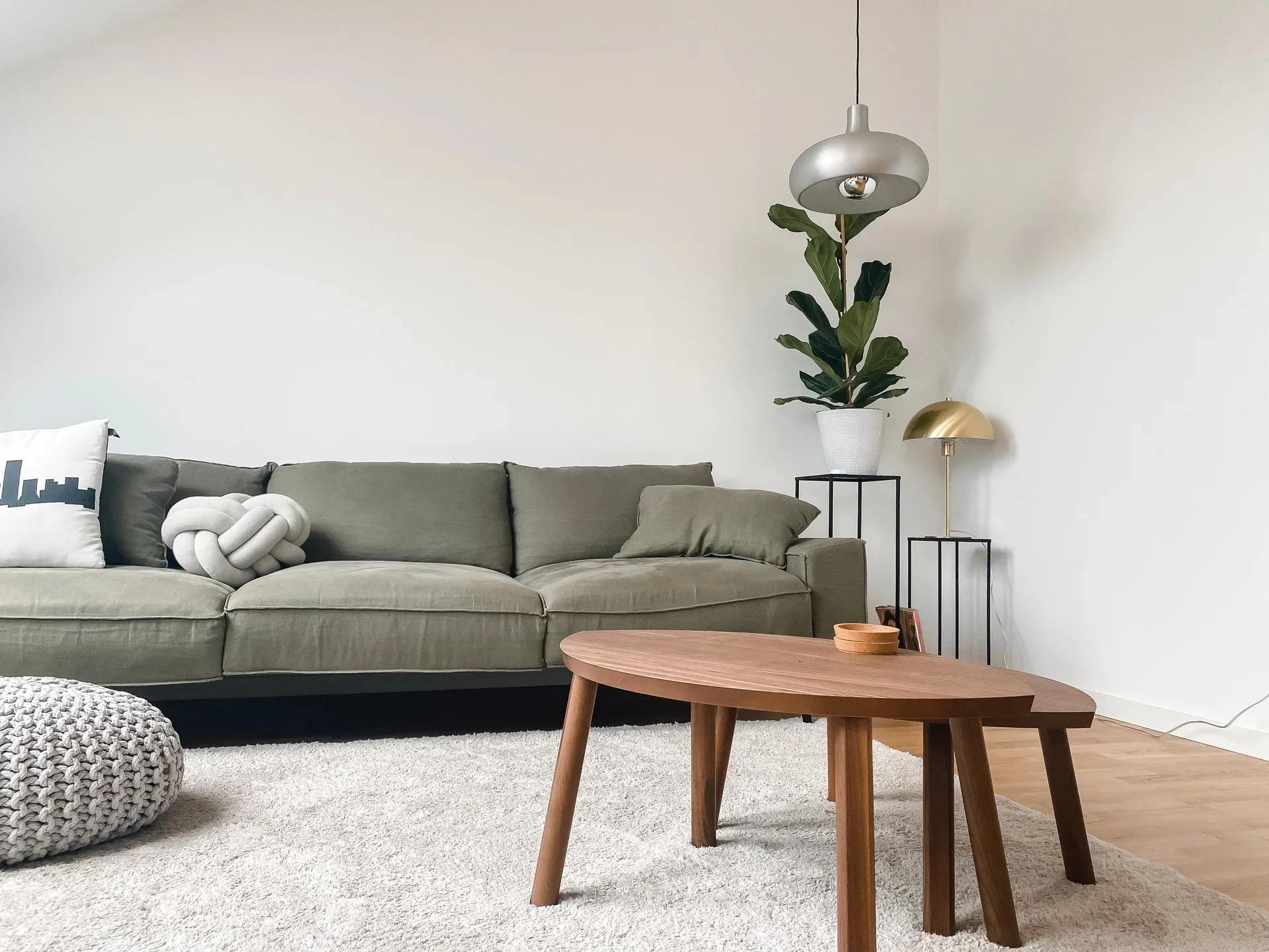 An olive green sofa sits on a plush white shag rug next to nested wooden coffee tables, a potted plant, and a metallic pendant lamp.