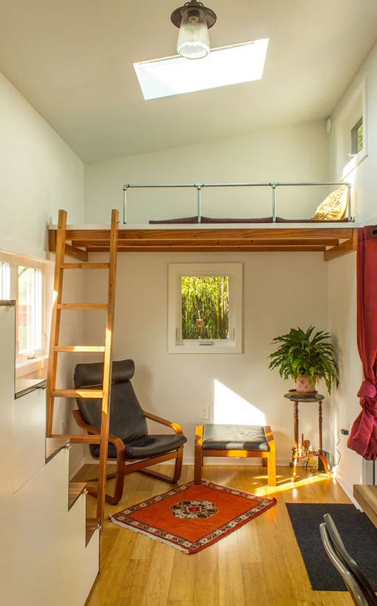 A tiny home interior featuring a wooden sleeping loft with a ladder, a black leather chair with an ottoman, and a small orange rug.
