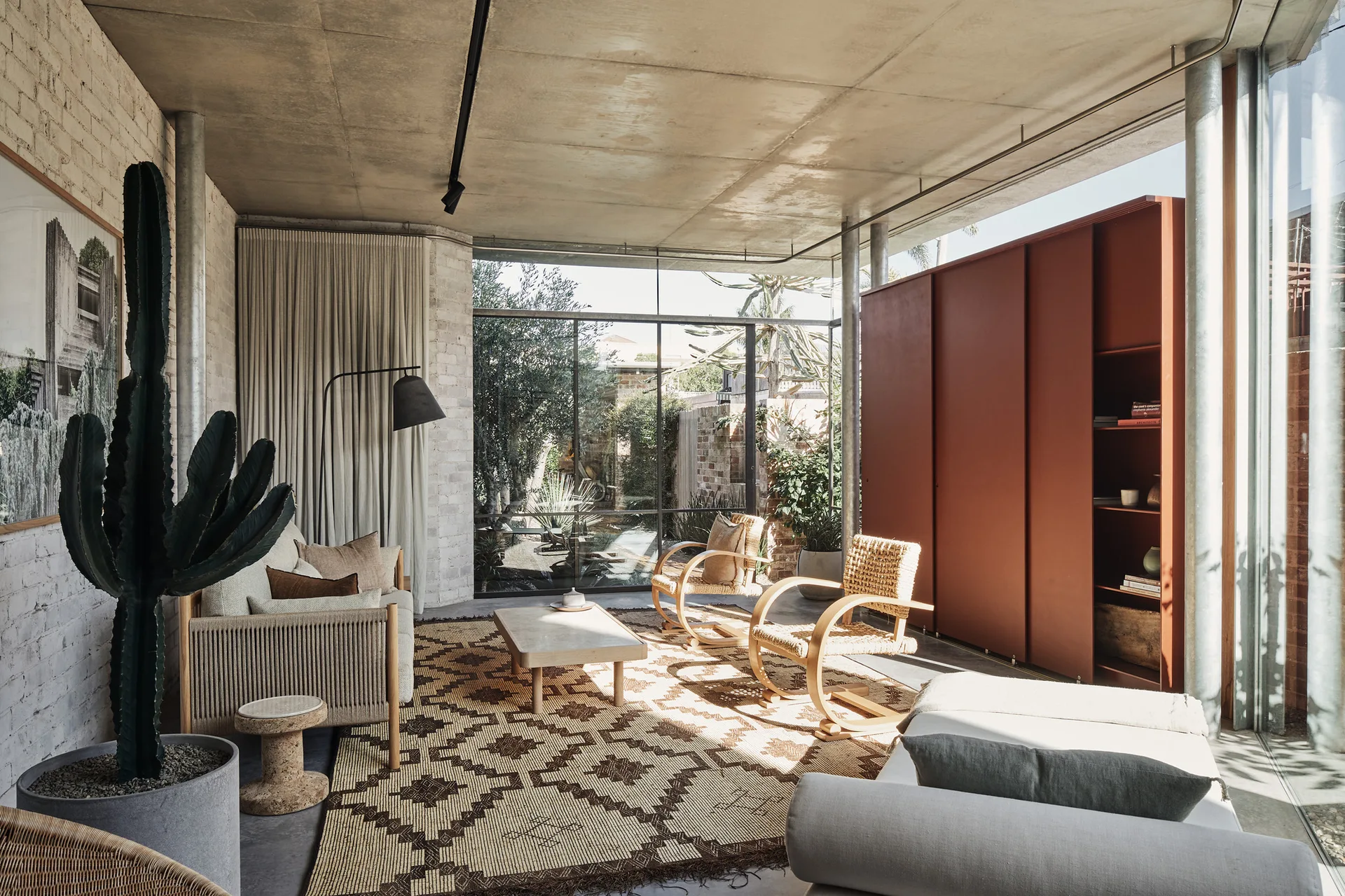 An industrial living room features a patterned geometric rug, two bentwood rattan armchairs, an orange cabinet, and an exposed concrete ceiling.