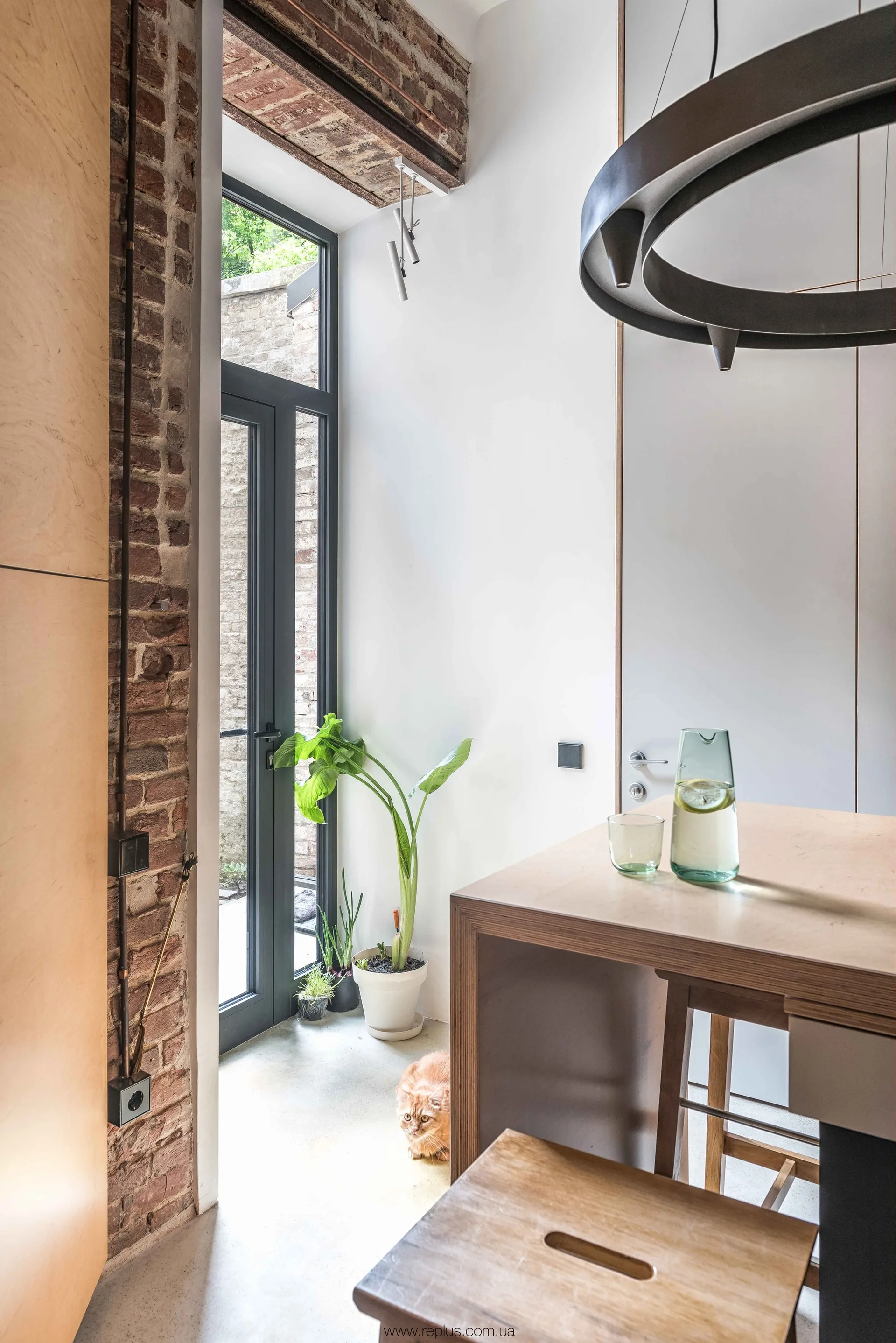 A modern kitchen features a wooden island, an exposed brick wall, a tall black-framed glass door, and a light grey concrete floor.