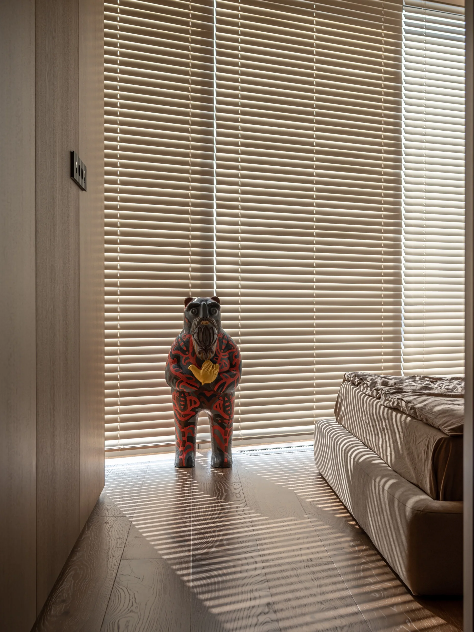 An art sculpture stands on a wooden floor in a bedroom corner next to a bed and a large window with venetian blinds.