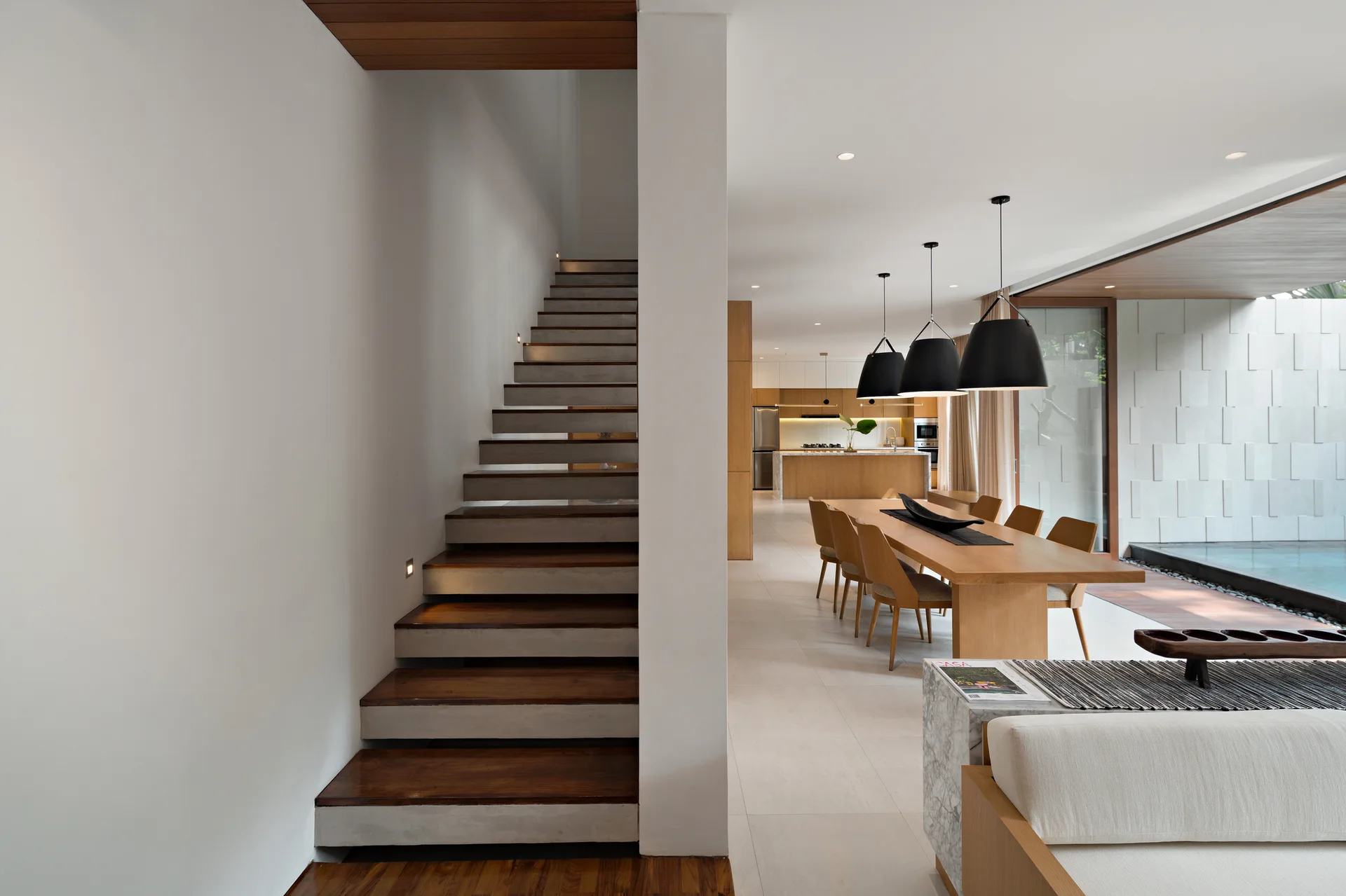 A modern home interior features a wooden staircase with recessed step lights next to an open dining area with black pendant lamps.