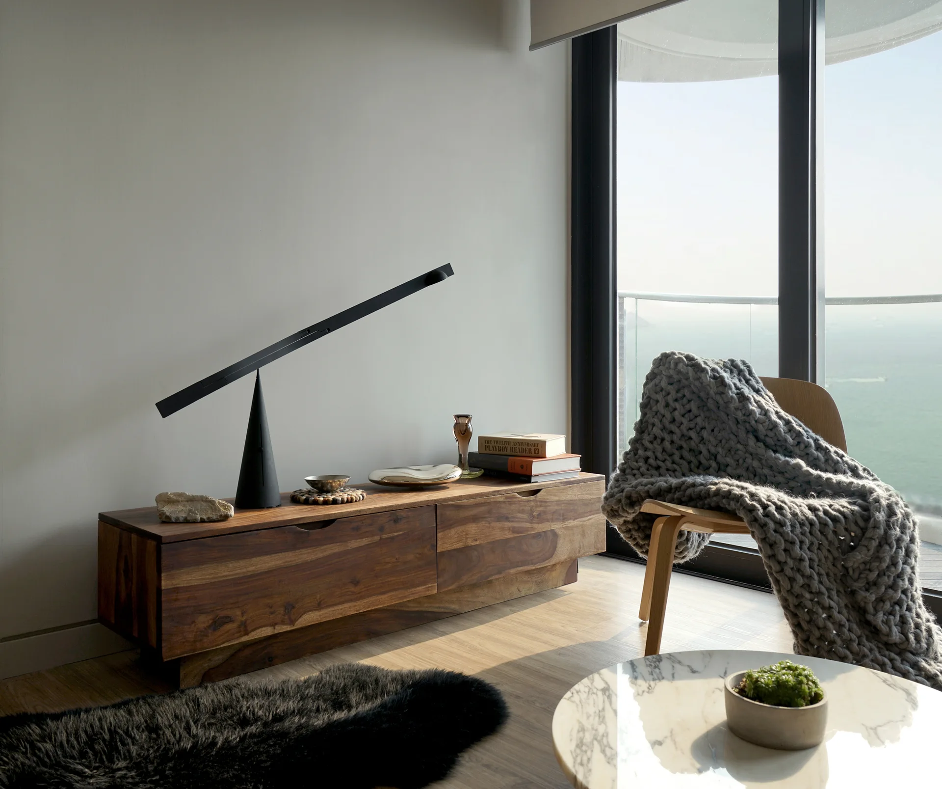 A low wooden media console holds a sculptural black cone lamp next to an armchair draped with a chunky grey knit blanket before a balcony overlooking the sea.