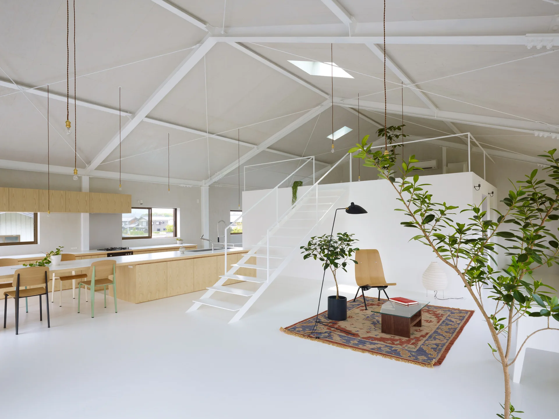 An open-plan minimalist living and kitchen space featuring a vaulted ceiling with white trusses, a light wood kitchen island, and a white open staircase.