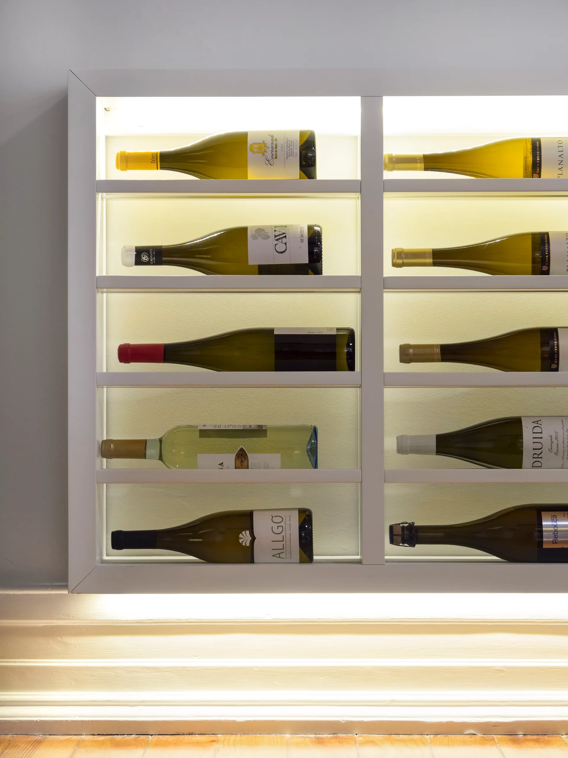 A modern built-in white wine rack with soft backlighting displaying wine bottles horizontally on a light wall above a classic baseboard.