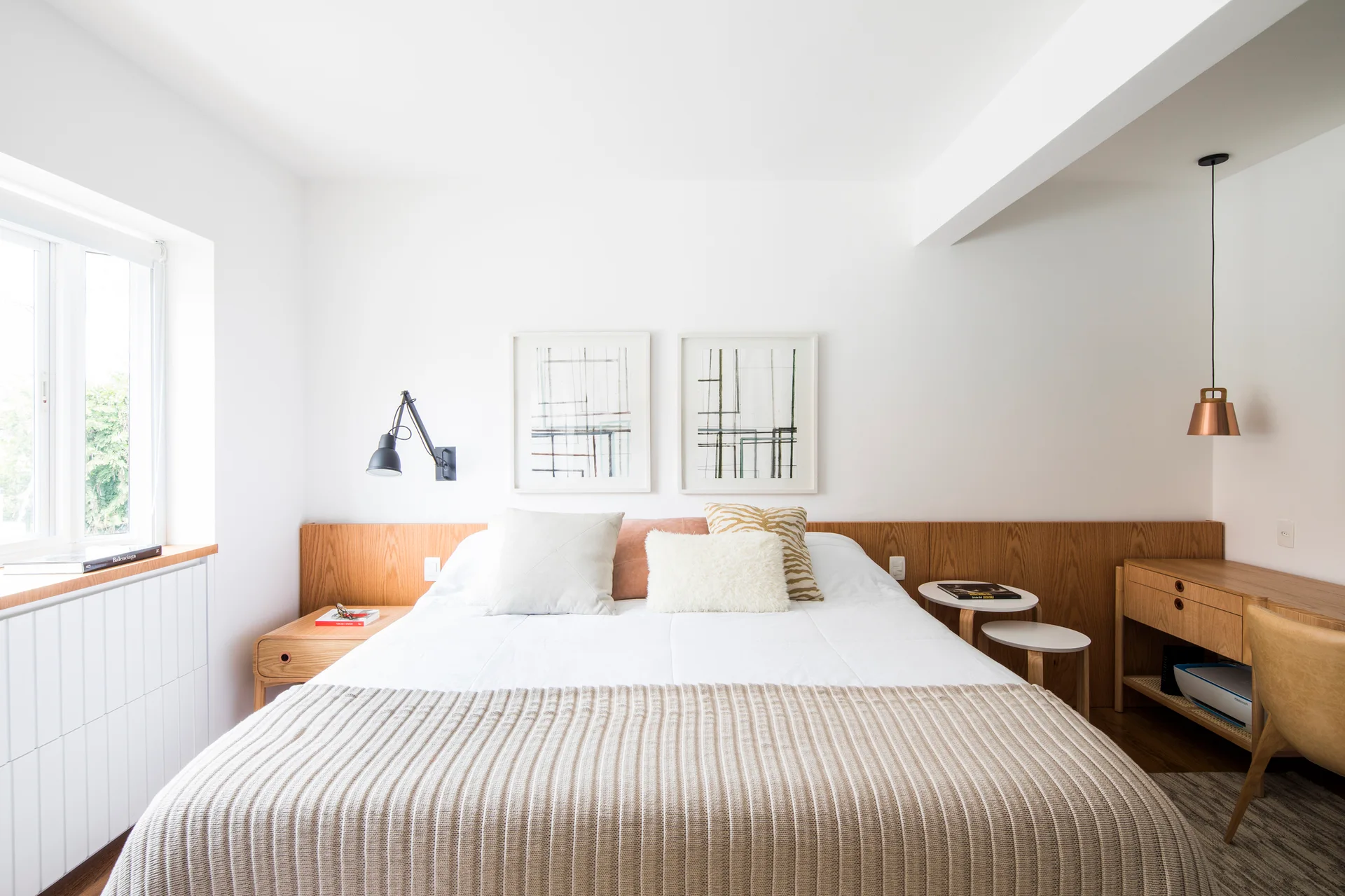 A minimalist bedroom features a bed with white bedding, a continuous low wood headboard panel, an integrated desk, and framed art.