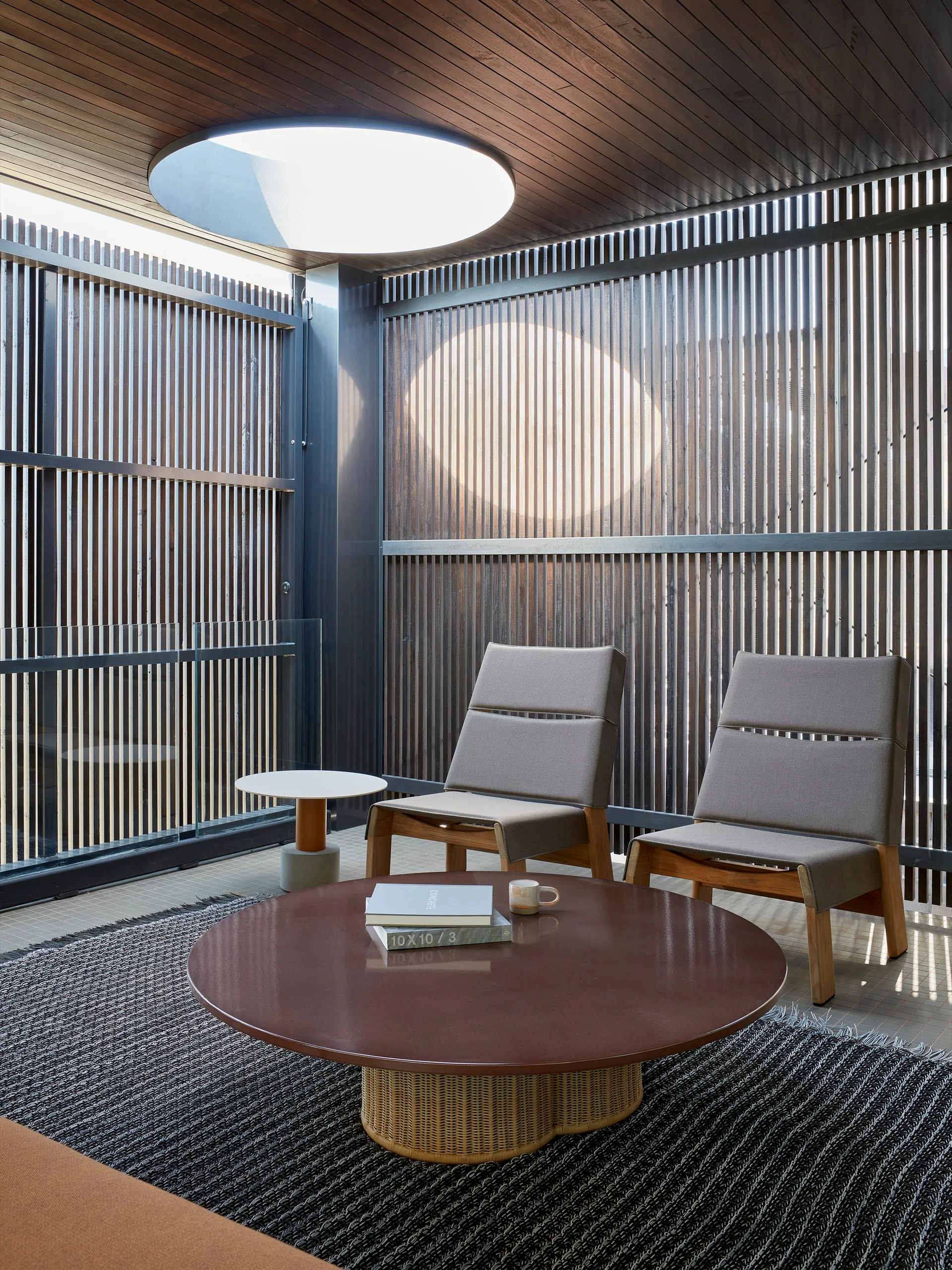 A contemporary seating area features two grey fabric armchairs, a round brown coffee table, and a vertical wood slat wall below a circular skylight.