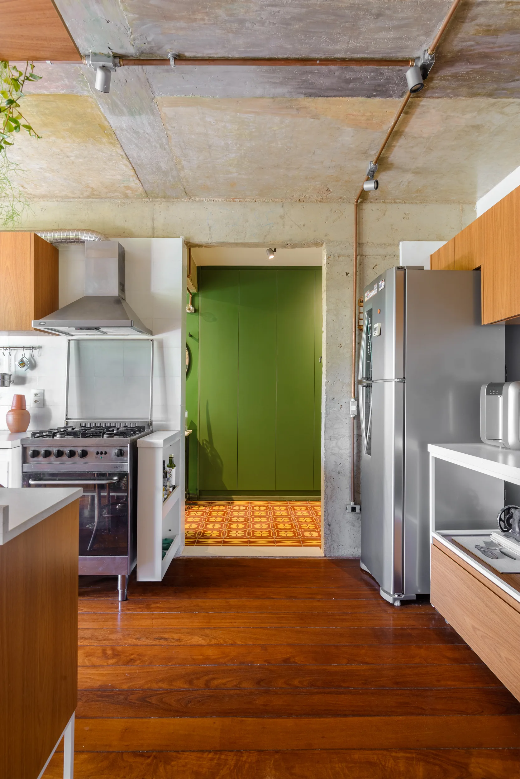 An industrial kitchen with an exposed concrete ceiling, stainless steel appliances, and a doorway leading to a green hallway with patterned tiles.