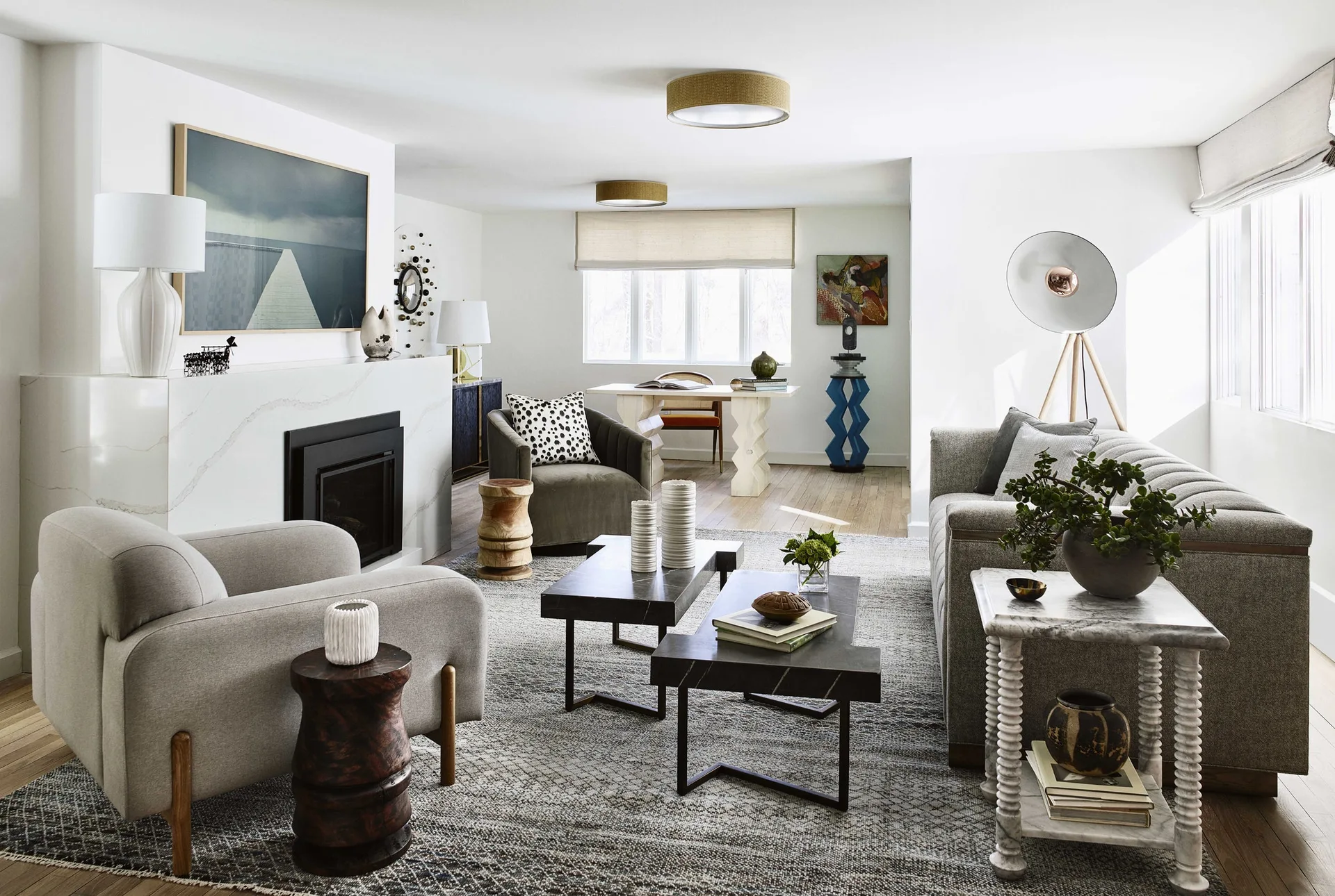 A modern living room features a white stone fireplace, grey upholstered armchairs, and two dark stone coffee tables on a large patterned rug.