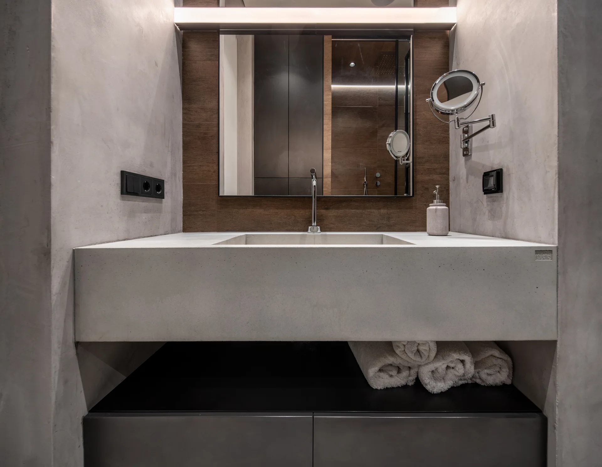 A modern bathroom vanity features a thick concrete basin, dark wood paneling behind a mirror, and concrete plaster walls.