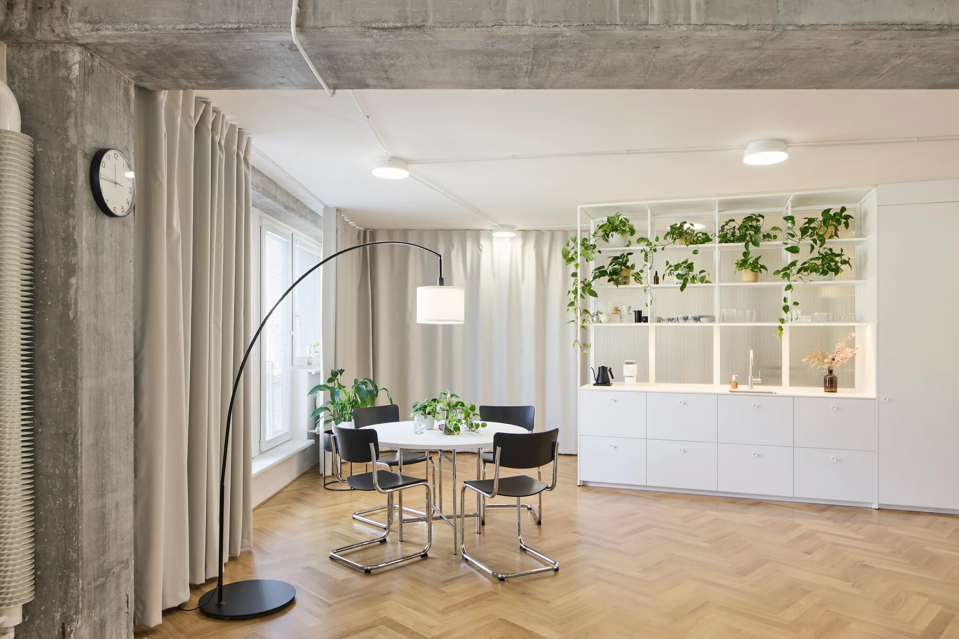 A minimalist dining area featuring a round white table, black metal-framed chairs, a white shelving unit, and an exposed concrete column.