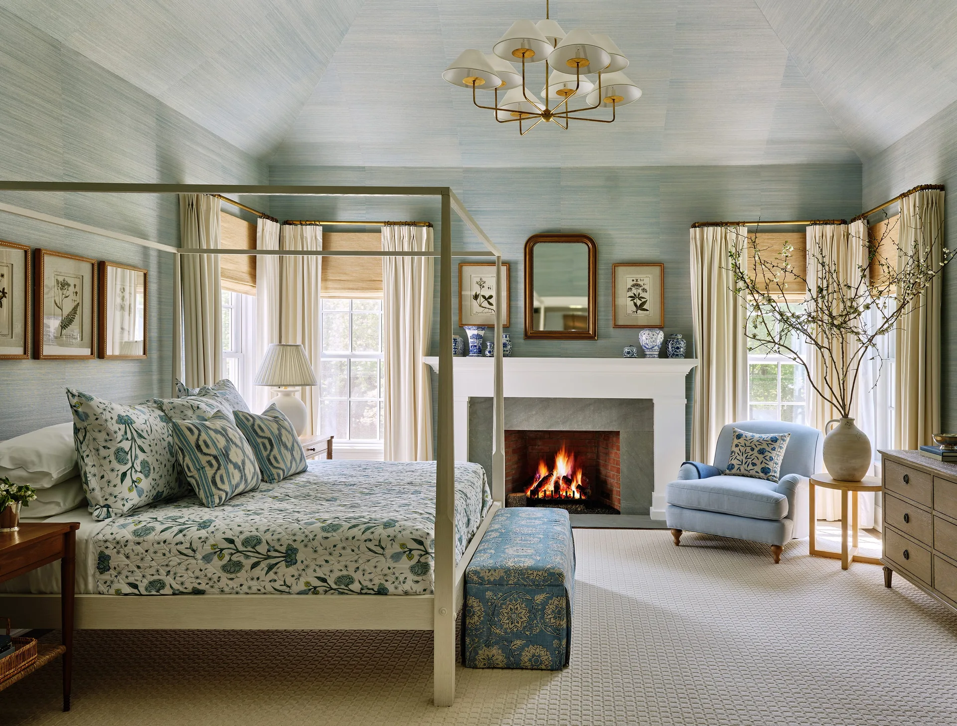 A bedroom with light blue grasscloth walls and a vaulted ceiling, featuring a four-poster bed, a brick fireplace, and an armchair.