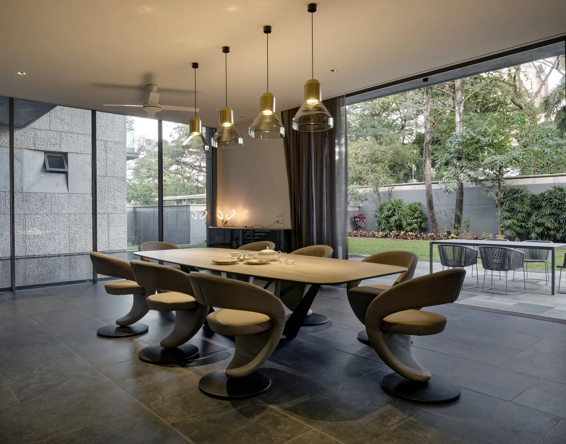 A modern dining room features sculptural beige chairs around a large table, lit by pendant lights, beside glass doors opening to an outdoor patio.