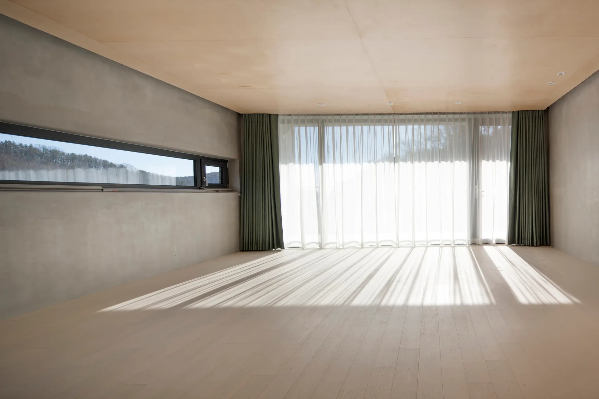 An empty room features grey concrete walls, a light wood floor, a plywood paneled ceiling, and large windows with sheer and green curtains.