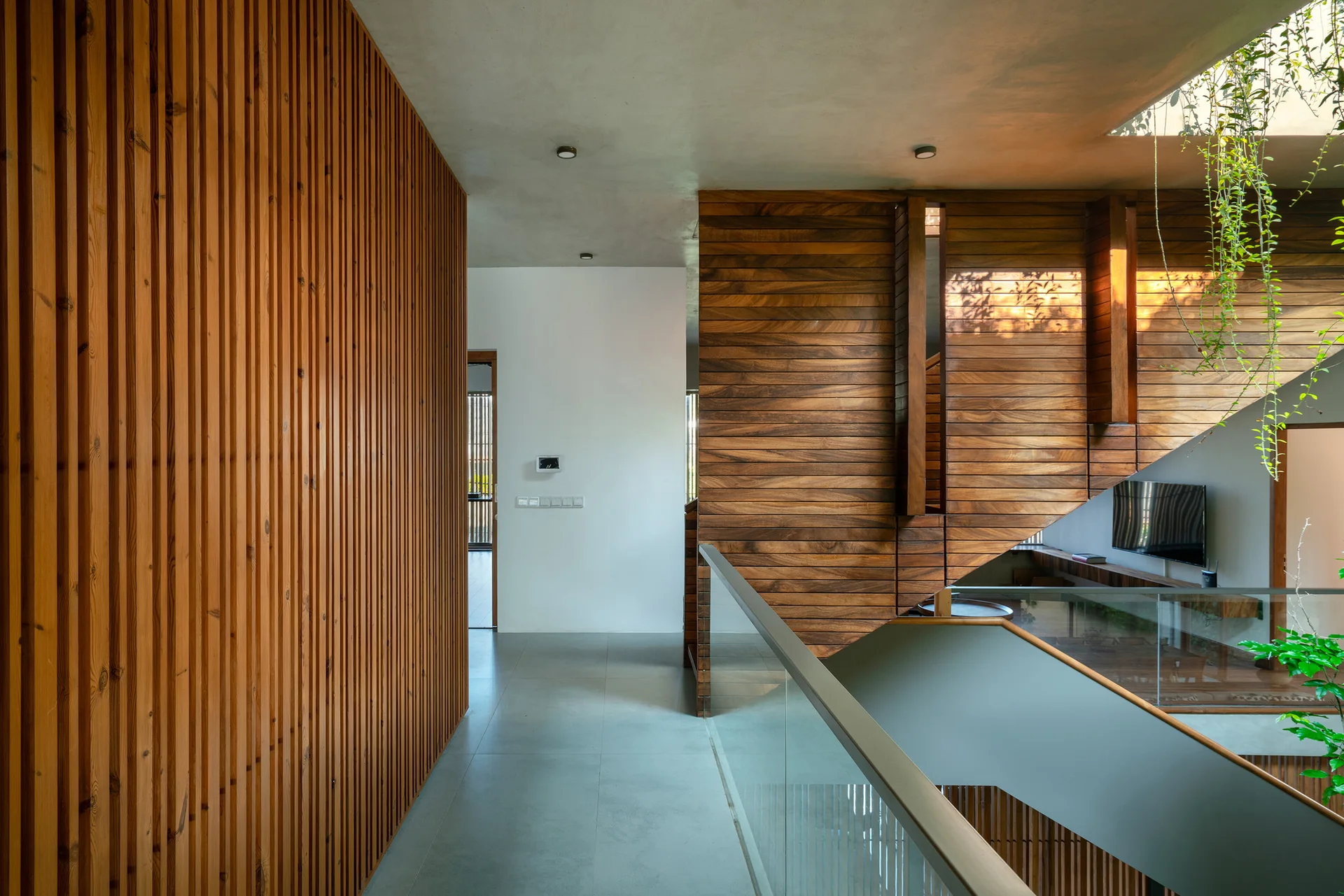 An interior hallway features a vertical slatted wood wall, a smooth concrete ceiling, and a glass-railed staircase with hanging green plants.
