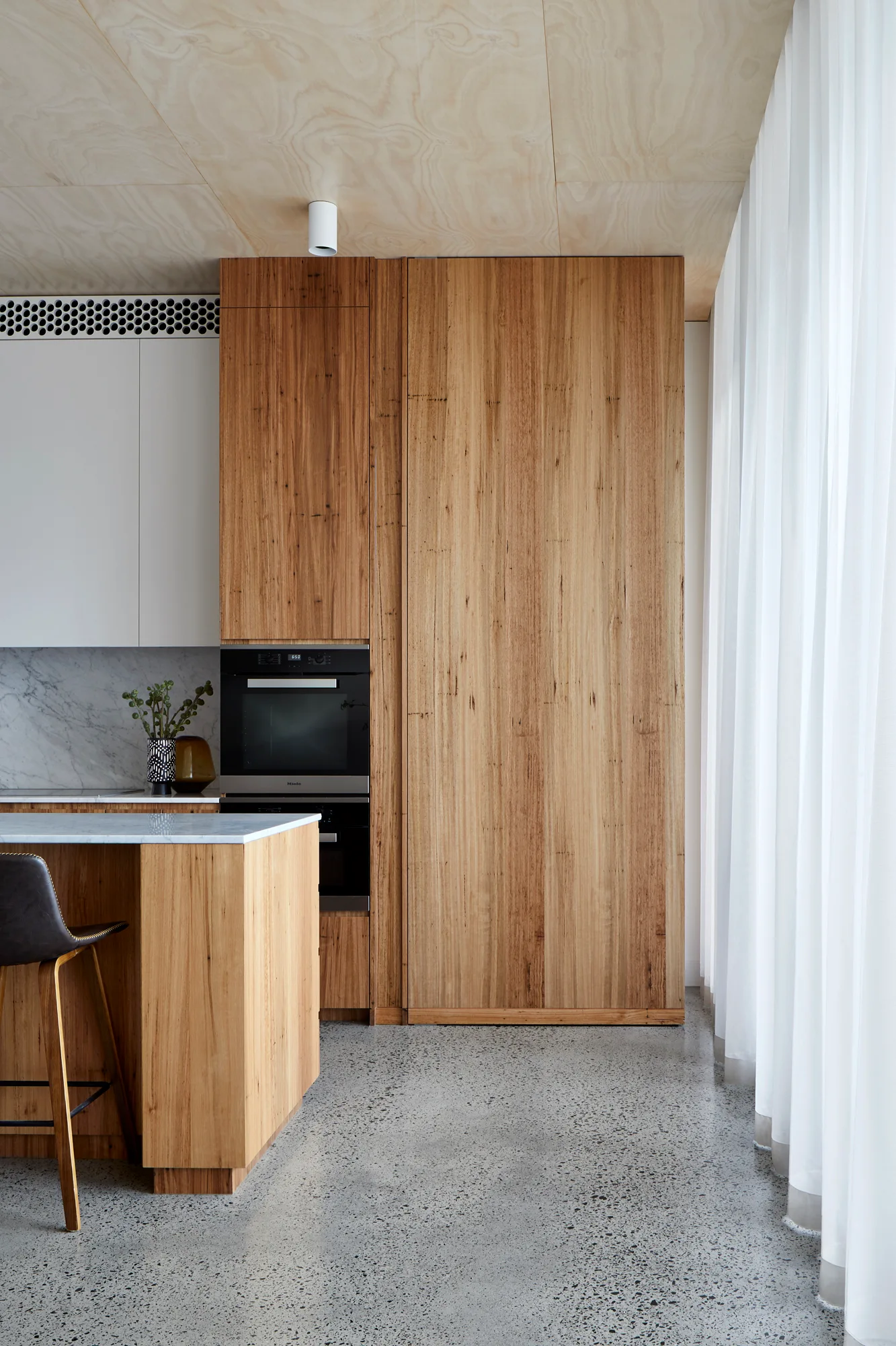 A minimalist kitchen features warm wood cabinetry, a plywood ceiling, and a polished concrete floor alongside a sheer white curtain.