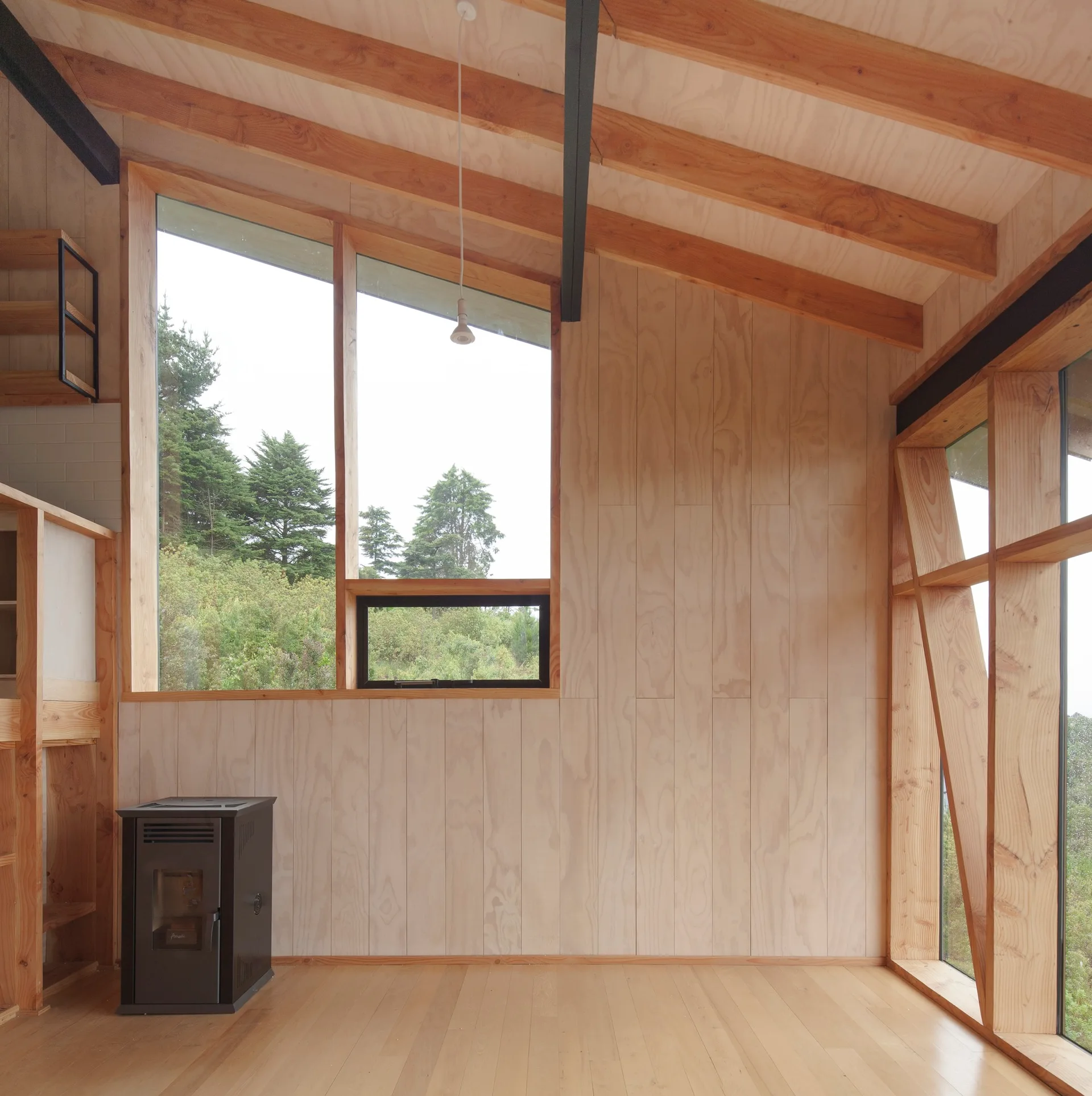 A cabin interior features light plywood walls, exposed ceiling rafters, large windows overlooking green trees, and a black wood-burning stove.