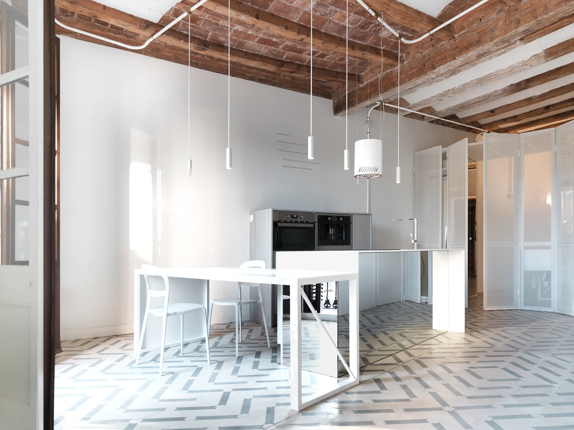 A modern white kitchen features a geometric patterned tile floor, minimalist island, slim white pendant lights, and an exposed brick and wood beam ceiling.