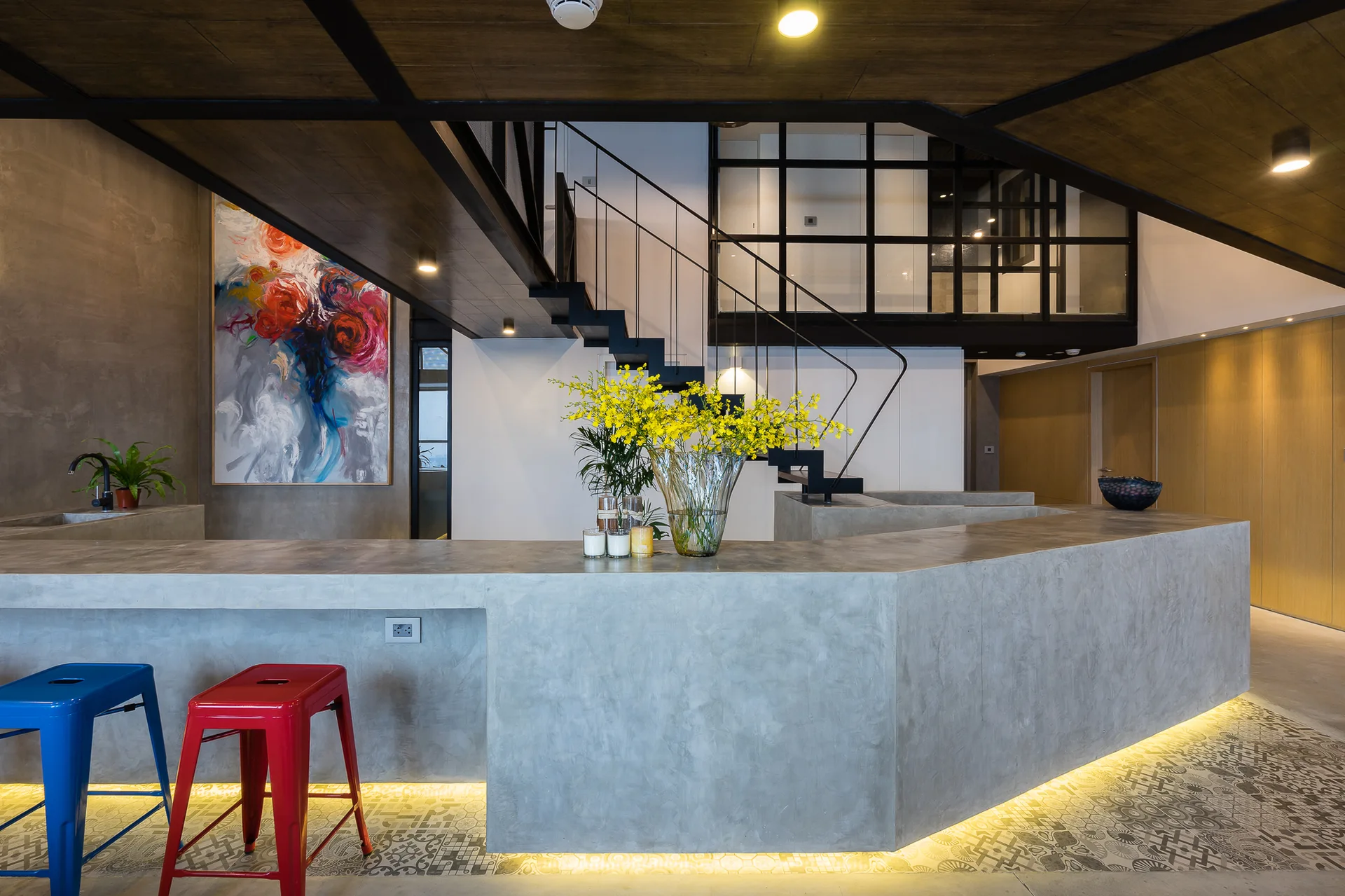 A modern open kitchen featuring a cast concrete island with red and blue metal bar stools, a metal staircase, and LED base lighting.